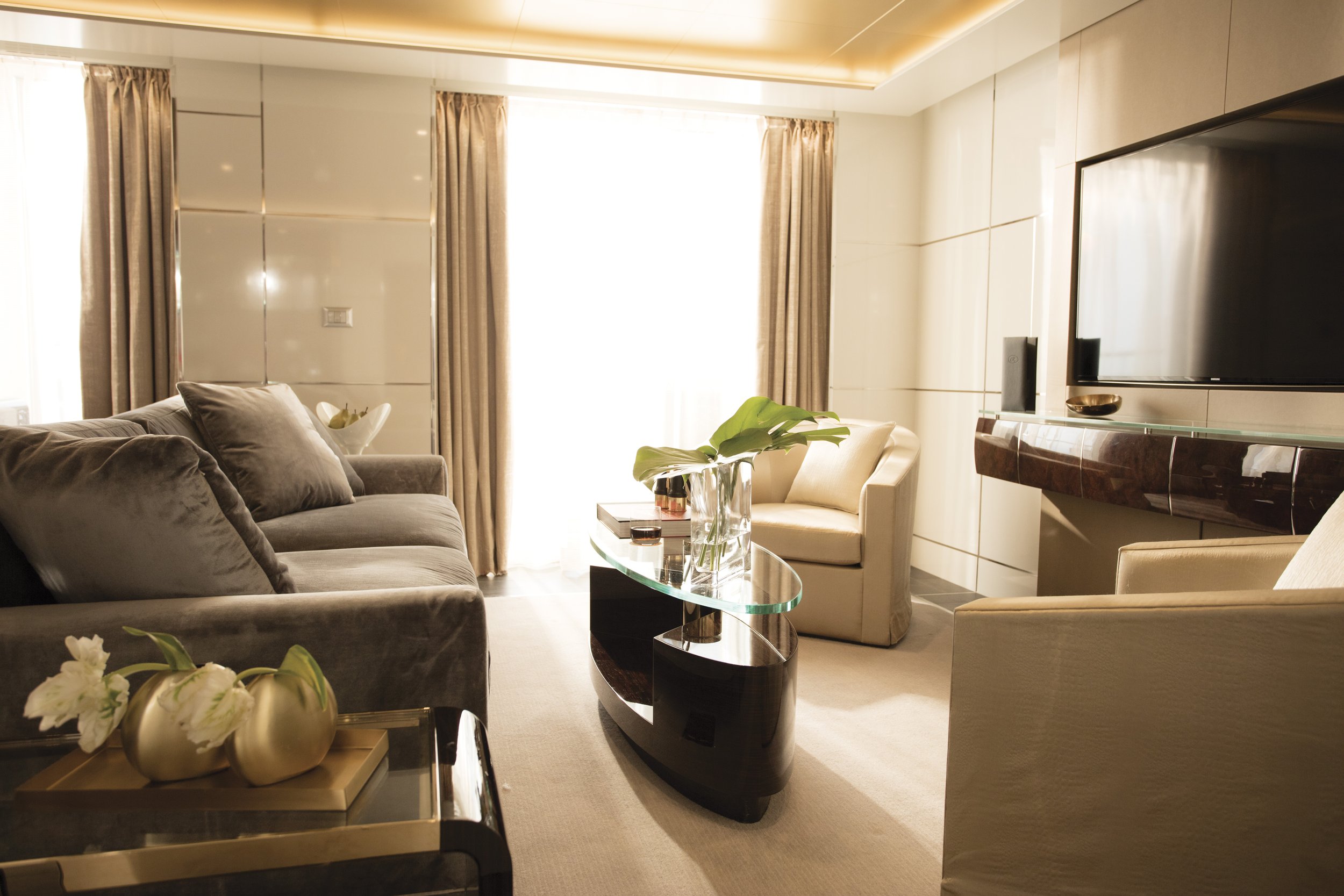 A modern living room with beige and gray furniture, including a sofa and armchair, a glass coffee table with a plant, and a wall-mounted flat-screen TV. There are large windows with beige curtains and a decorative bowl on a glass console. A vase with white flowers is in the foreground.