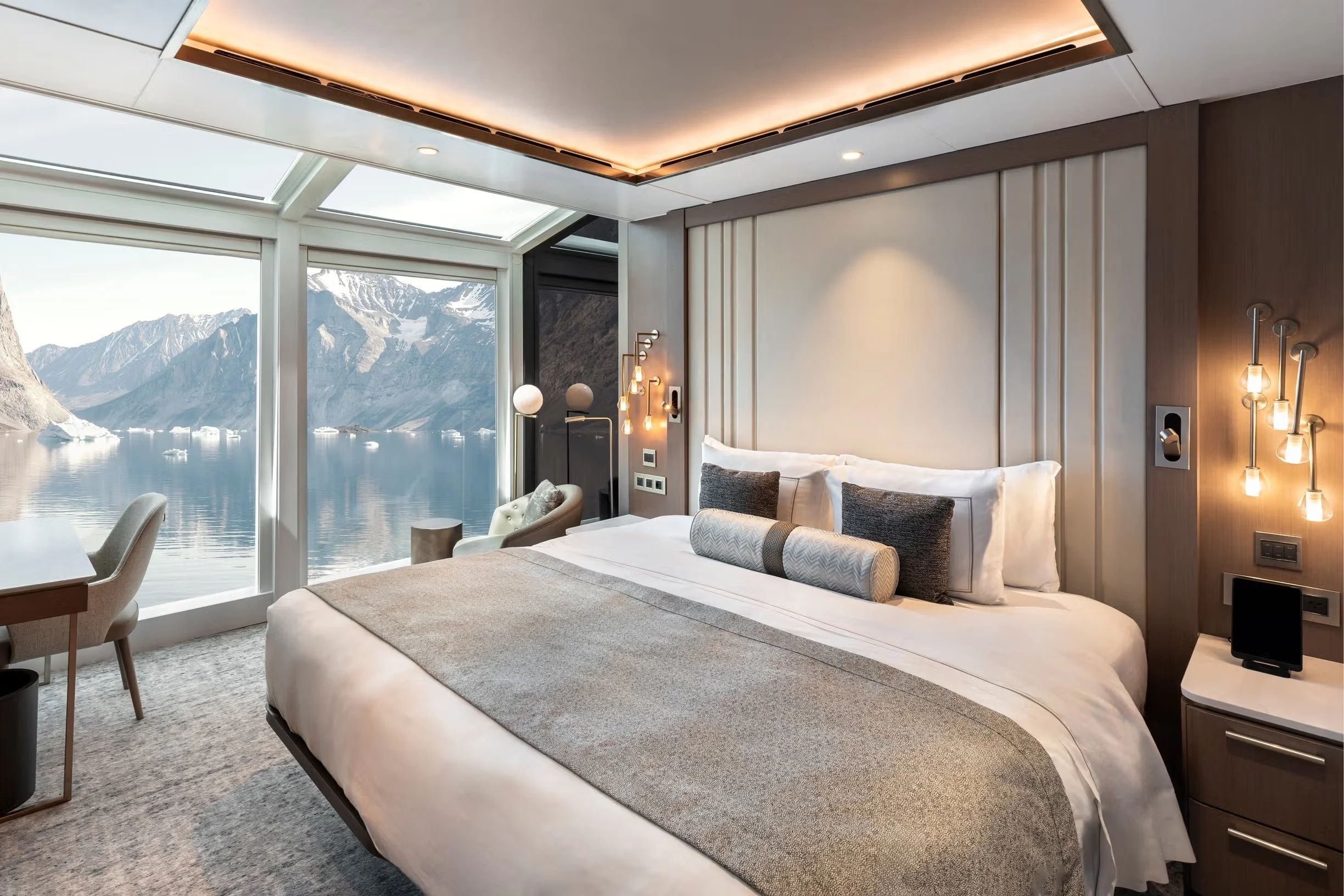 Elegant hotel room with a large bed, modern lighting fixtures, and a panoramic window overlooking snowy mountains and a body of water.
