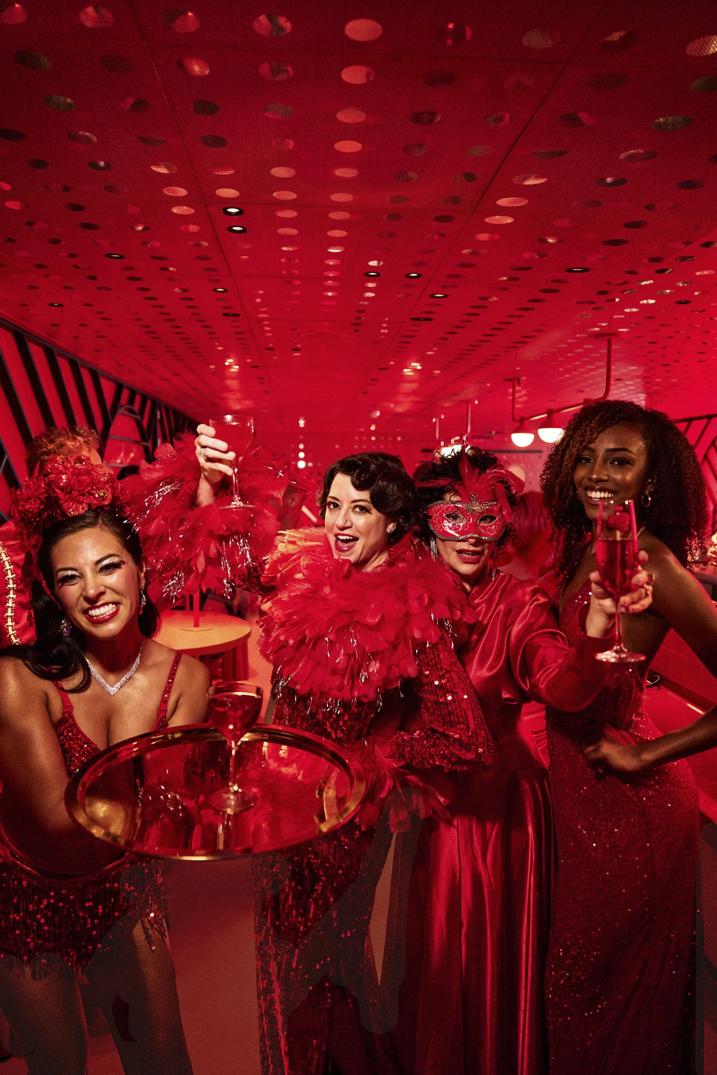 A group of women dressed in red at a festive celebration, holding glasses with red drinks, some wearing red feathers and masks, in a red-lit room.