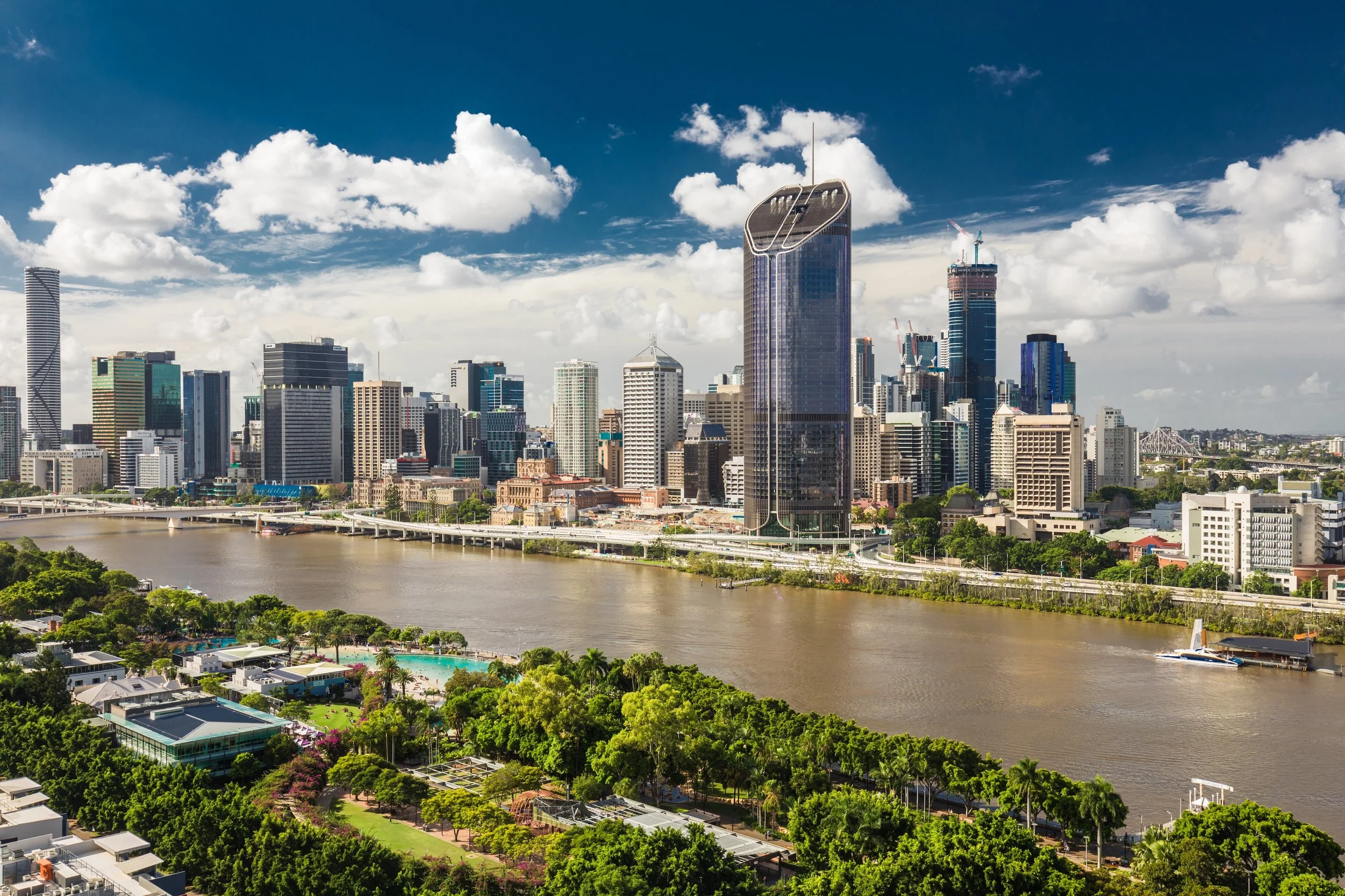 A city skyline featuring tall modern skyscrapers with some under construction, a river in the foreground, and a partly cloudy blue sky.