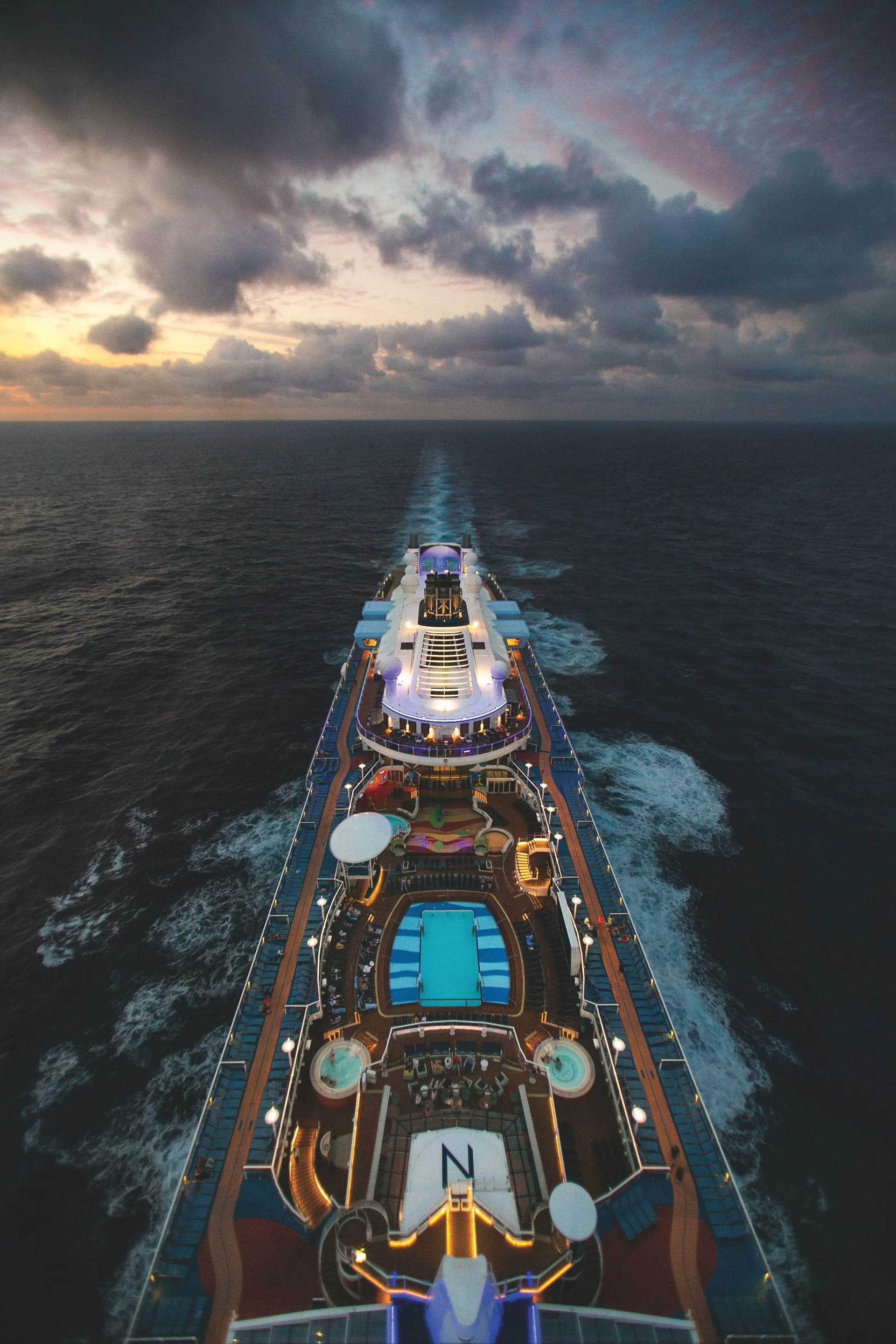 Top view of a cruise ship sailing through the ocean during sunset, with a cloudy sky overhead.
