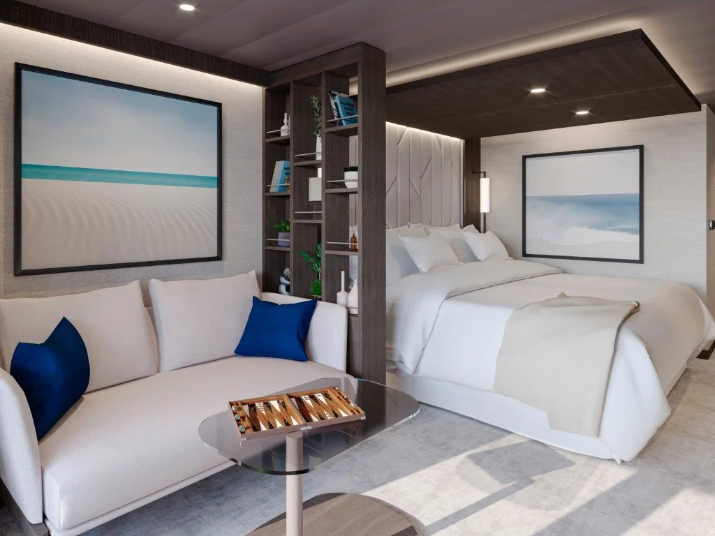 Modern hotel room with a large bed, white bedding, and artwork of a beach scene on the wall. There is a white sofa with blue pillows, a small table with a decorative tray, and a built-in bookshelf between the sleeping area and sofa. The room has neutral tones with dark wood accents and subtle lighting.