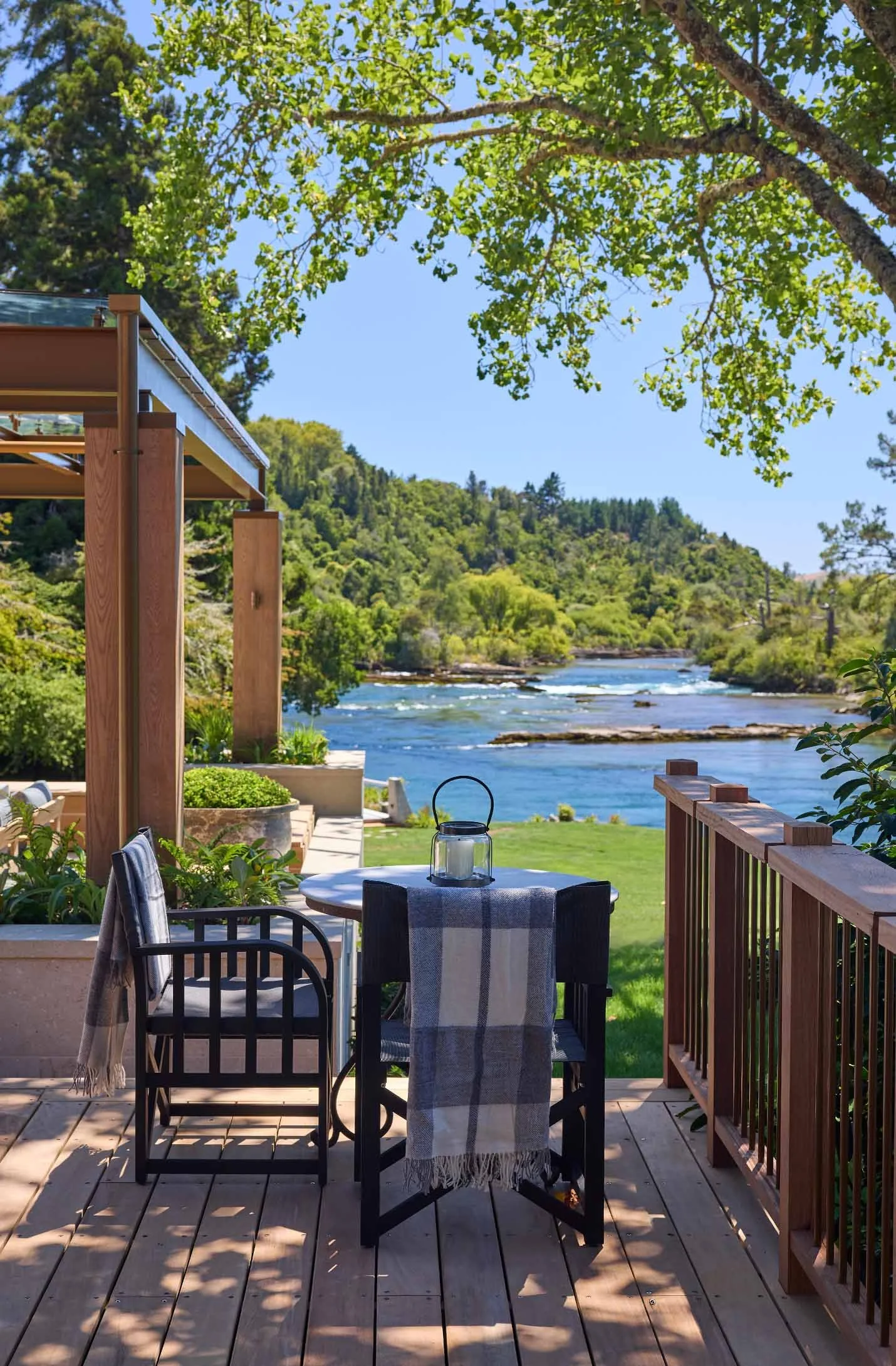 A wooden deck overlooking a river, with four chairs around a table draped with a striped cloth, a lantern on the table, and lush green trees and hills in the background under a clear blue sky.
