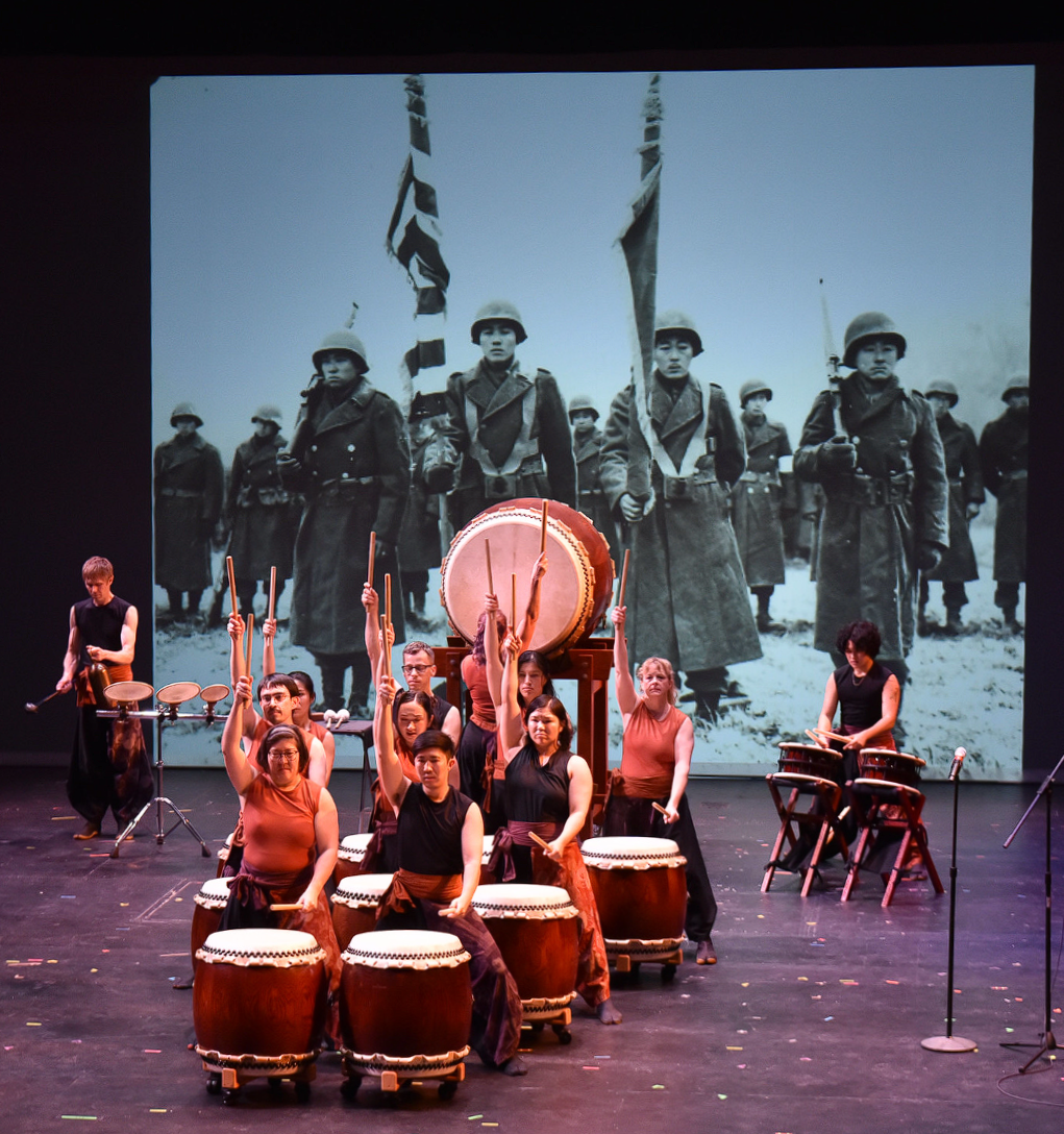 Taiko drummers with sticks raised stand in front of a black and white photo of the all-Japanese American WWII unit the 442nd.  Credit: Katharine Govea Saunders