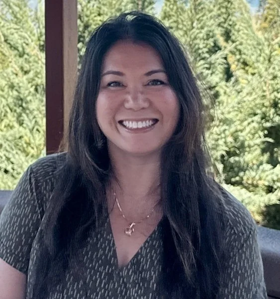 Lena, a woman with long dark hair smiles. Evergreen trees are in the background.