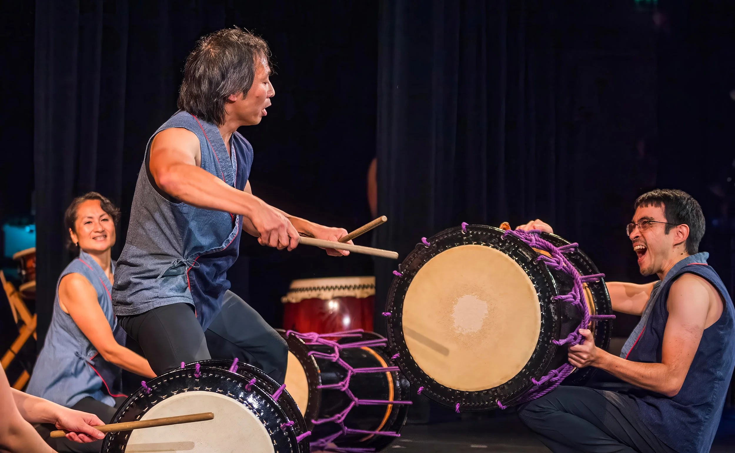 A kneeling taiko drummer holds a drum while another drummer prepares to strike it.