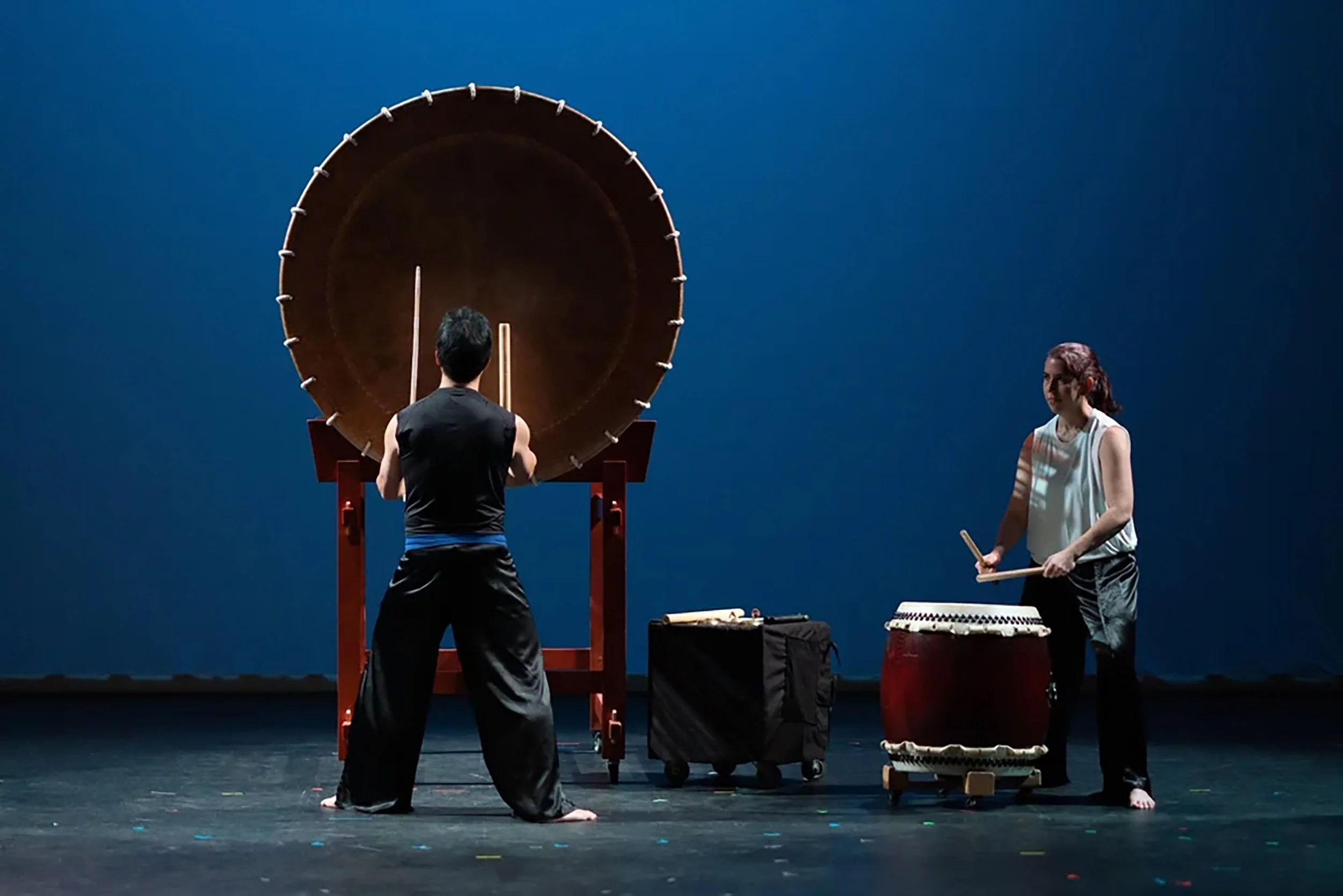 A drummer stands in front of a large drum with his back to the crowd. Another drummer plays a barrel-shaped drum stage left.