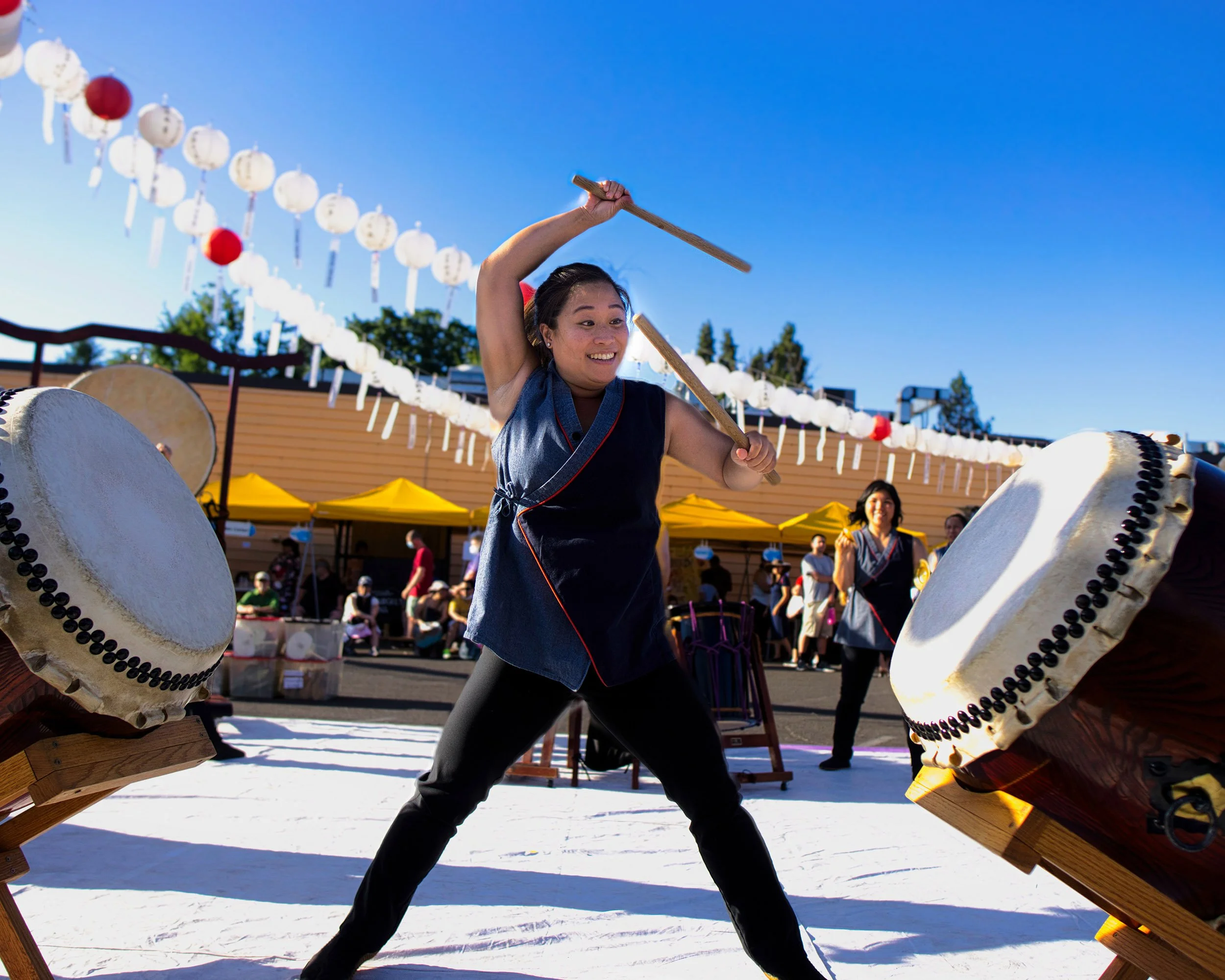 A taiko drummer stands between two drums on slanted stands. She waves a stick above her head and raises the other in front of her face. Above her hang red and white obon festival lanterns.