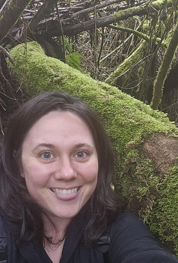 Kegan poses in front of a log in the forest. They smile with their tongue sticking out