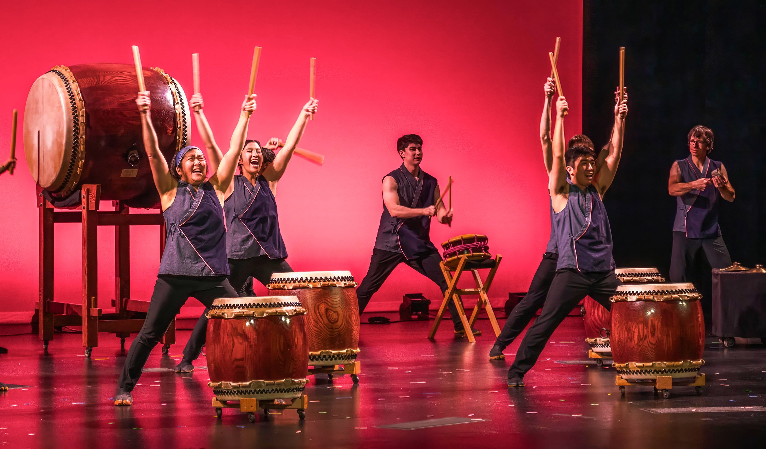 Four taiko drummers stand in front of a vivid, pinkish red backdrop with their arms raised. A fifth drummer keeps the beat in the background and a sixth performer plays a small, hand instrument.