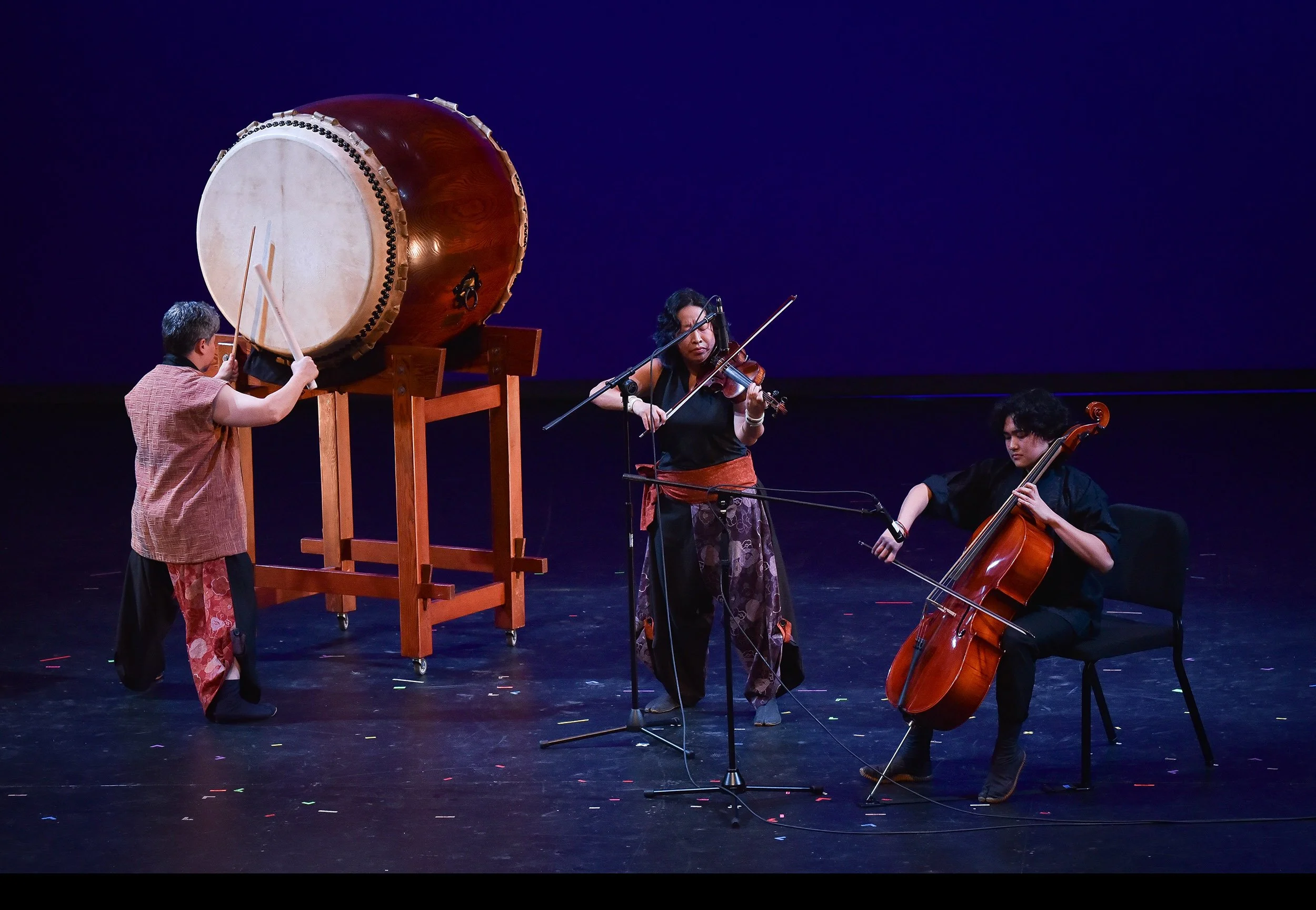 On a semi-dark stage, a violin player stands between a cello player and a taiko drummer. The taiko drummer, Tiffany Tamaribuchi, stands in front of a very large drum (odaiko).