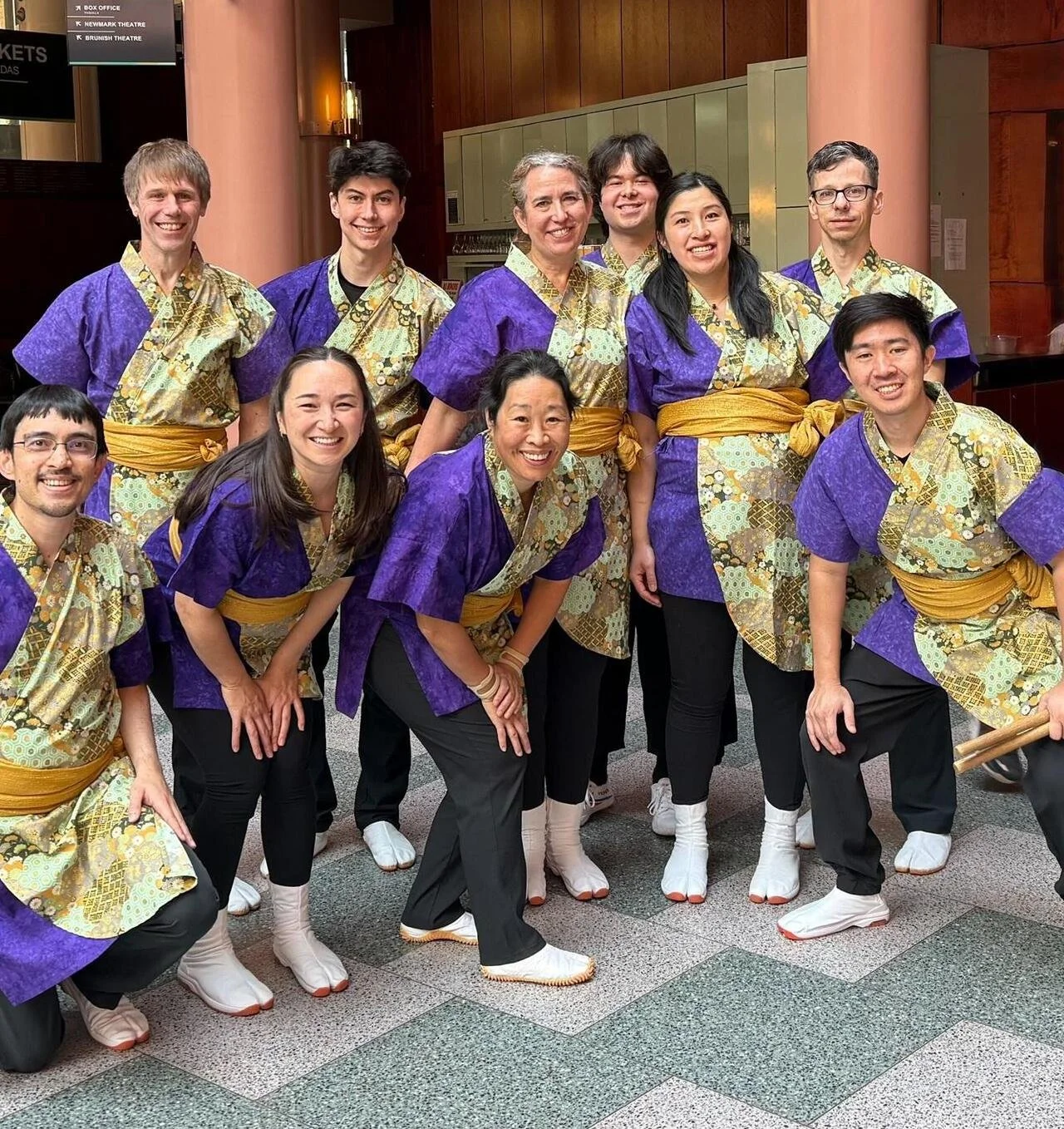 10 smiling Portland Taiko performers pose for a group shot wearing the new purple and gold costume