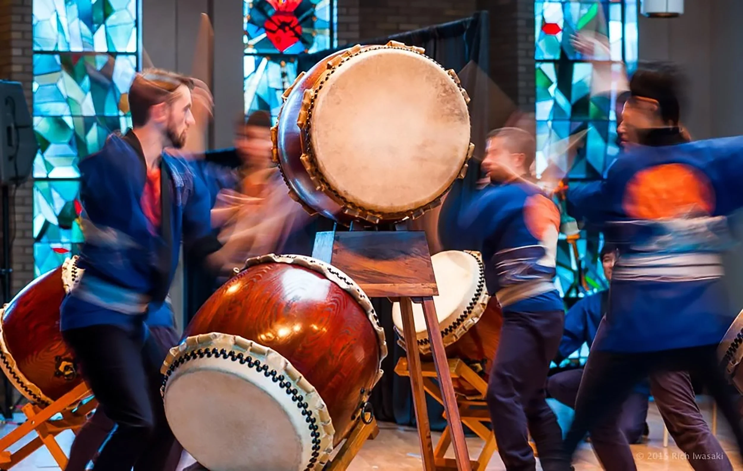 Four taiko drummers circle a large drum on a stand. They are moving so fast, they are blurry.