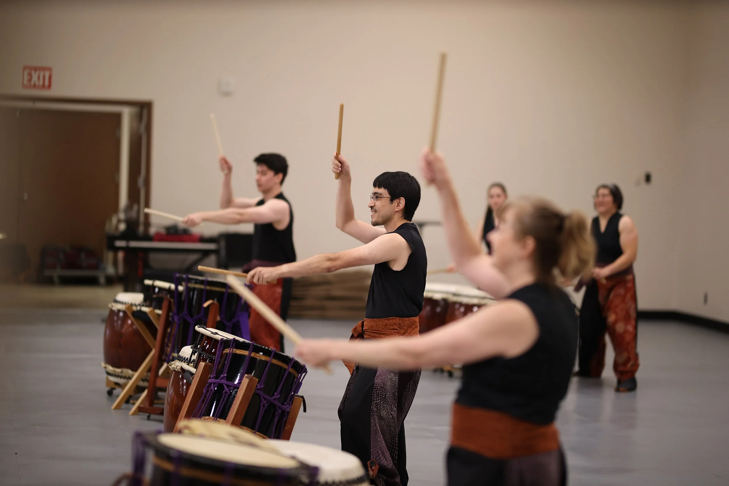 Five Portland Taiko performers at a local school. Three drummers have their sticks raised while two others pay instruments in the background.