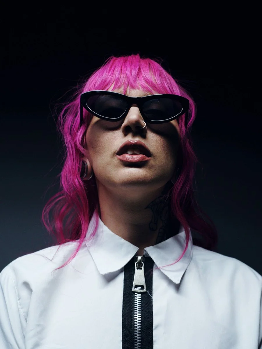 A person with pink hair wearing black sunglasses, a white zip-up jacket, and jewelry, against a dark background.