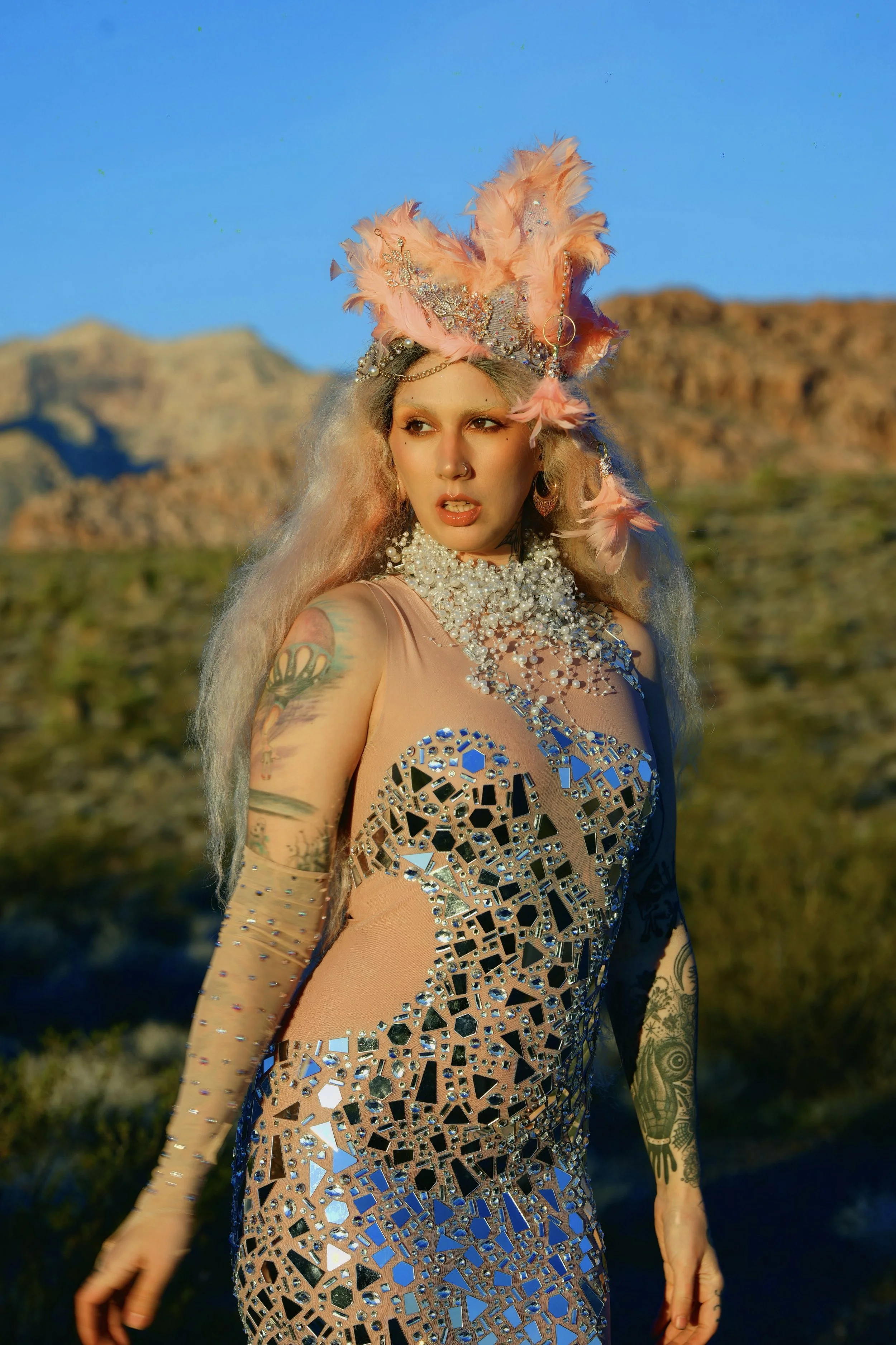 A woman with long, blonde hair wearing an elaborate, feathered headdress and a dress covered in mirrored pieces, standing outdoors in a desert landscape with mountains in the background during daytime.