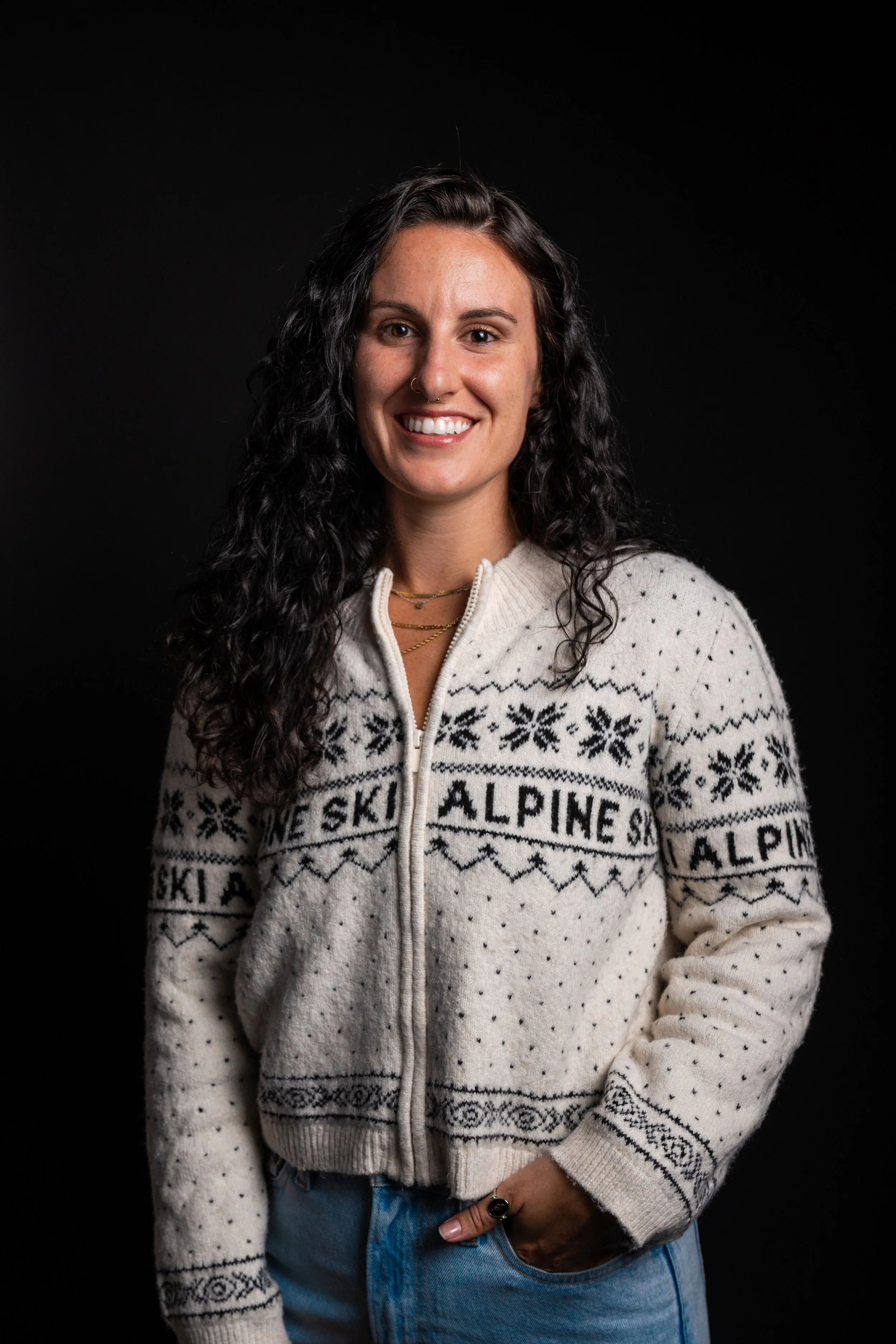 Woman with black hair smiling, wearing a white sweater and jeans against a dark backdrop.