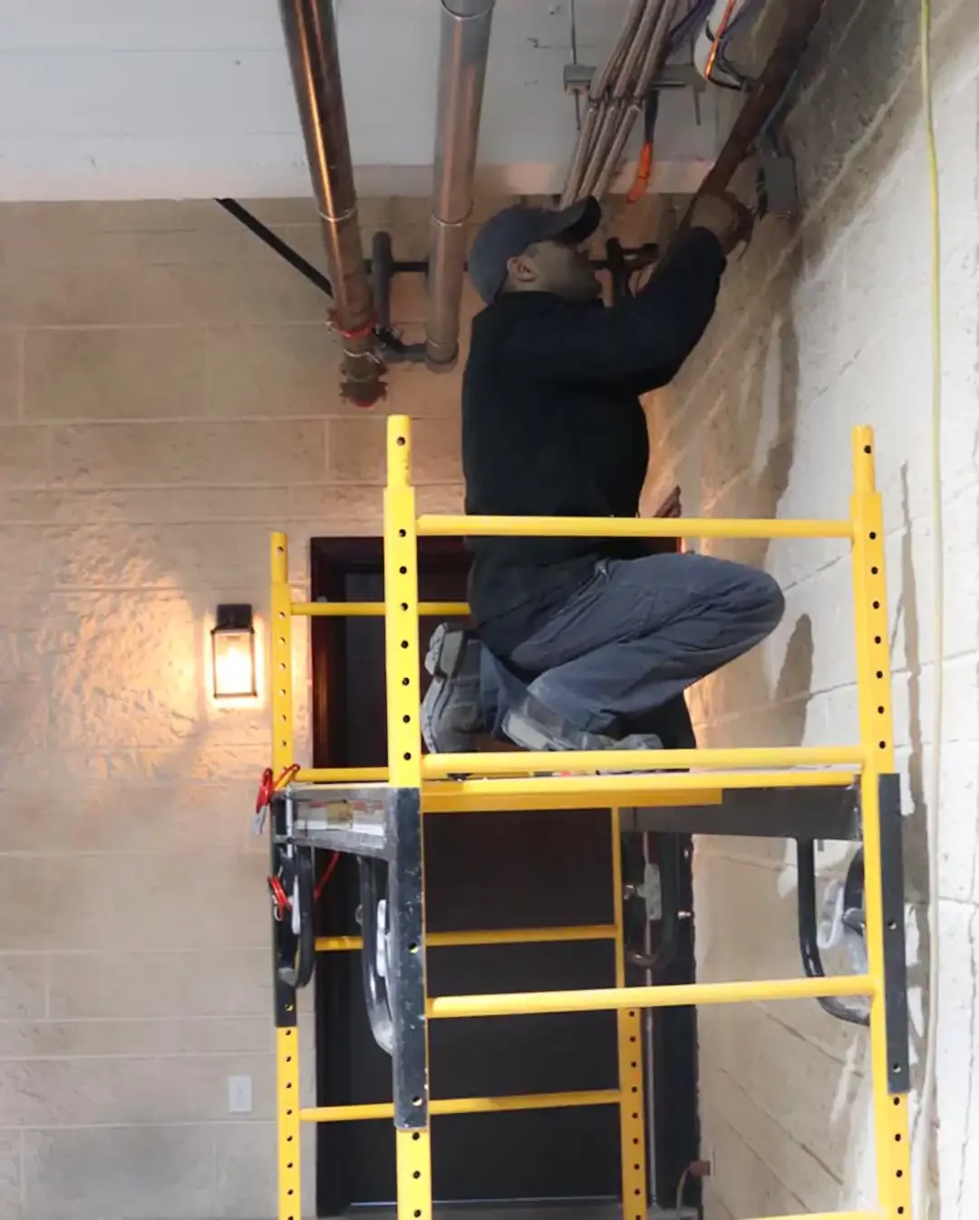 A professional HVAC technician working from a yellow scaffold to complete complex electrical wiring and piping for a commercial climate control system in a stone-walled facility.