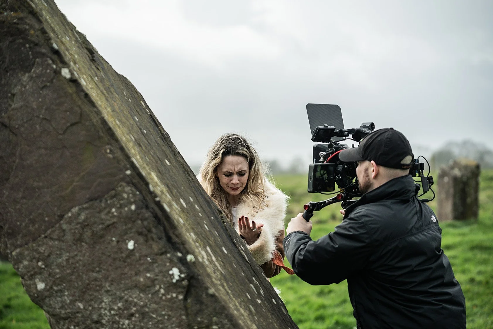A woman with long, curly blonde hair, wearing a white fur coat, is crying while being filmed outdoors on a cloudy day by a man with glasses and a black cap operating a professional camera.
