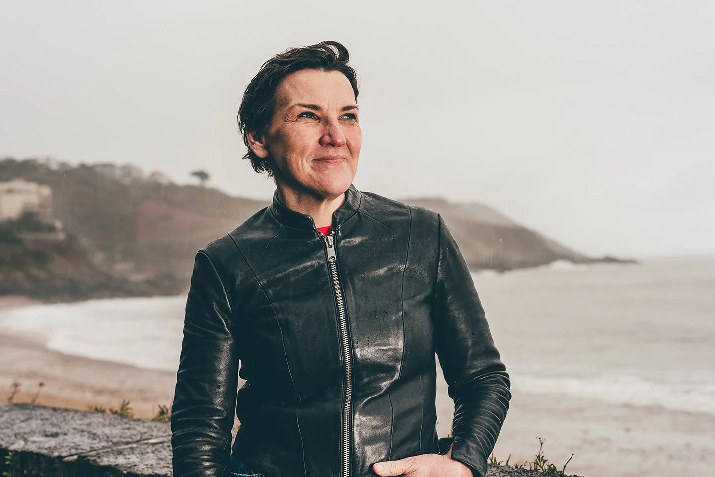 A woman with short dark hair wearing a black leather jacket standing on a beach with ocean and hills in the background.