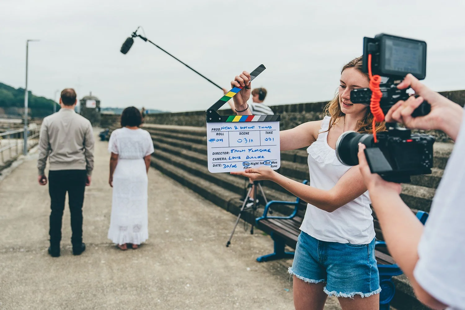 A film crew shooting an outdoor scene with three actors, a man, woman, and another man, standing on a path near a stone wall, with a woman holding a clapperboard to signal the start of filming.