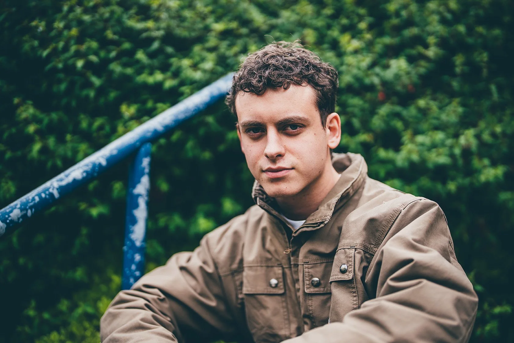 A young man with curly hair and a serious expression, wearing a brown jacket, sitting outdoors near green foliage.