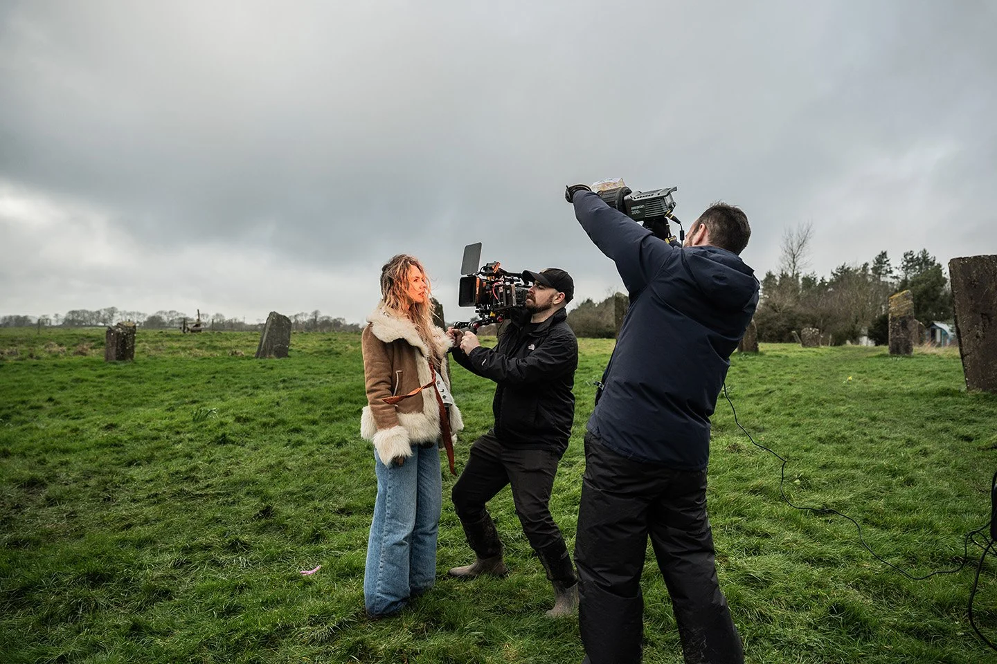 Filming scene outdoors with a woman being filmed by two cameramen on a grassy field with stone monuments and a cloudy sky.