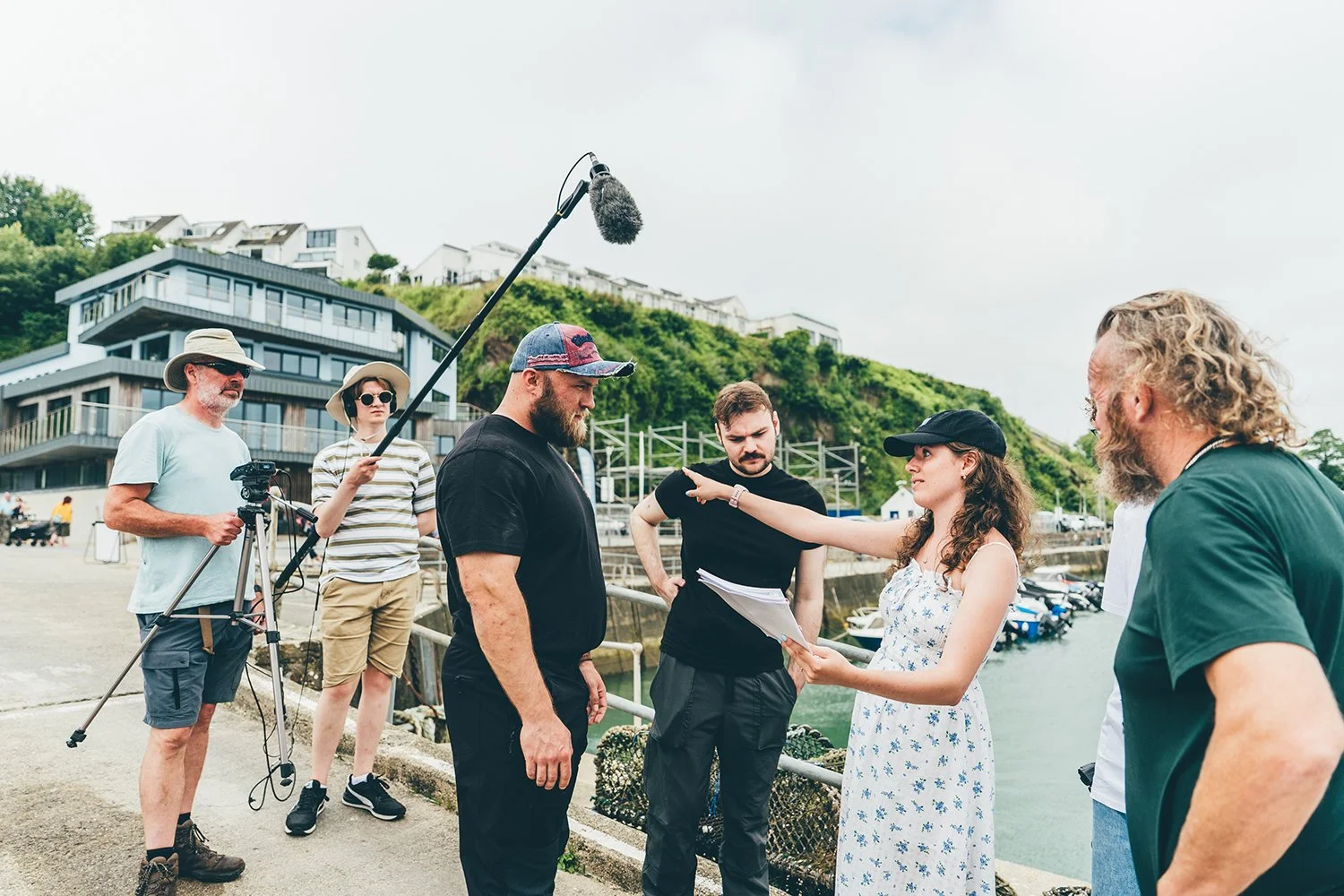 Filming crew on a waterfront with hills and modern houses, with a woman directing and holding a script, surrounded by camera operators and sound technicians.