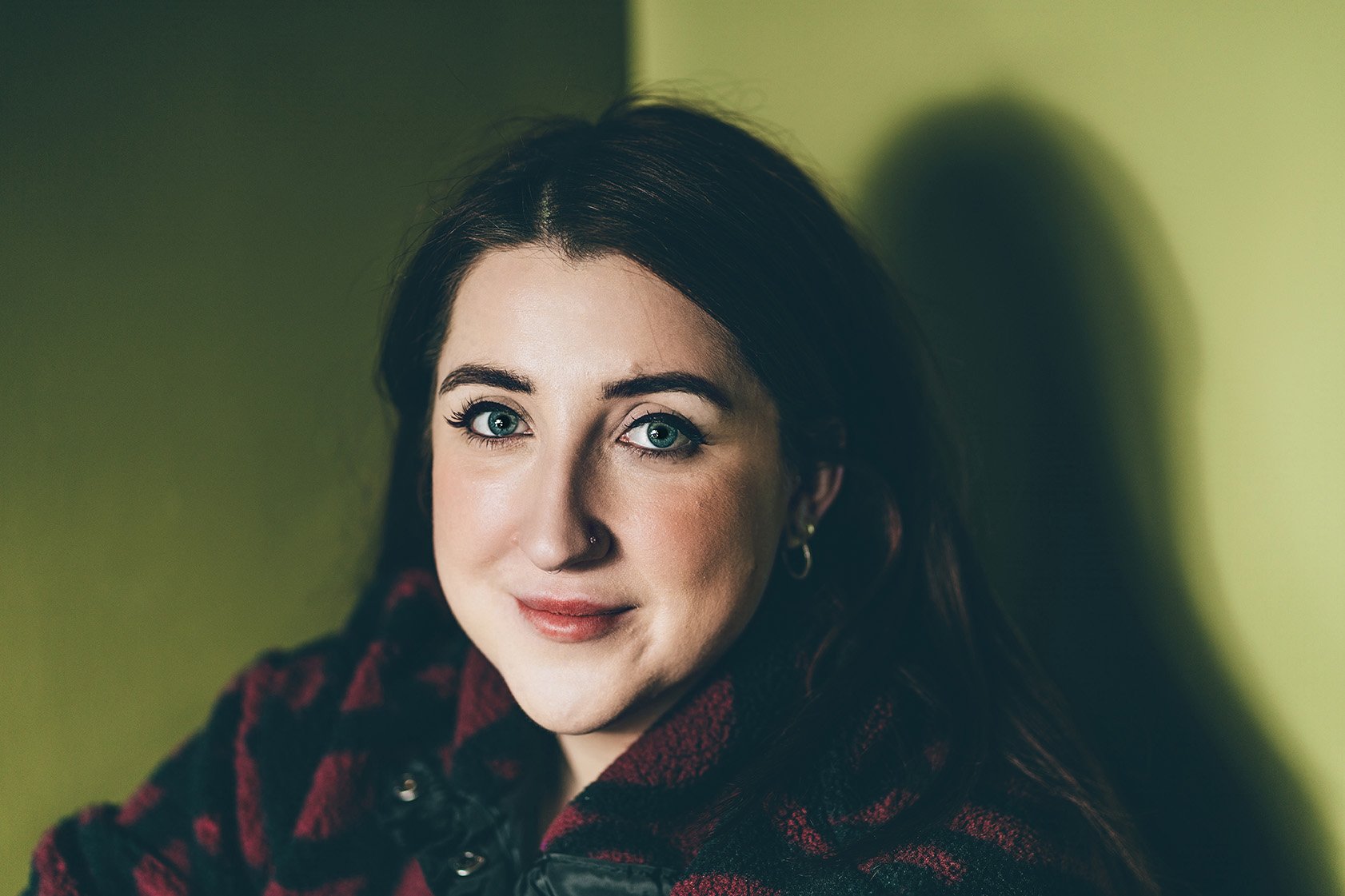 A young woman with dark hair, blue eyes, and a nose piercing, wearing a red and black plaid shirt, smiling softly against a green background.