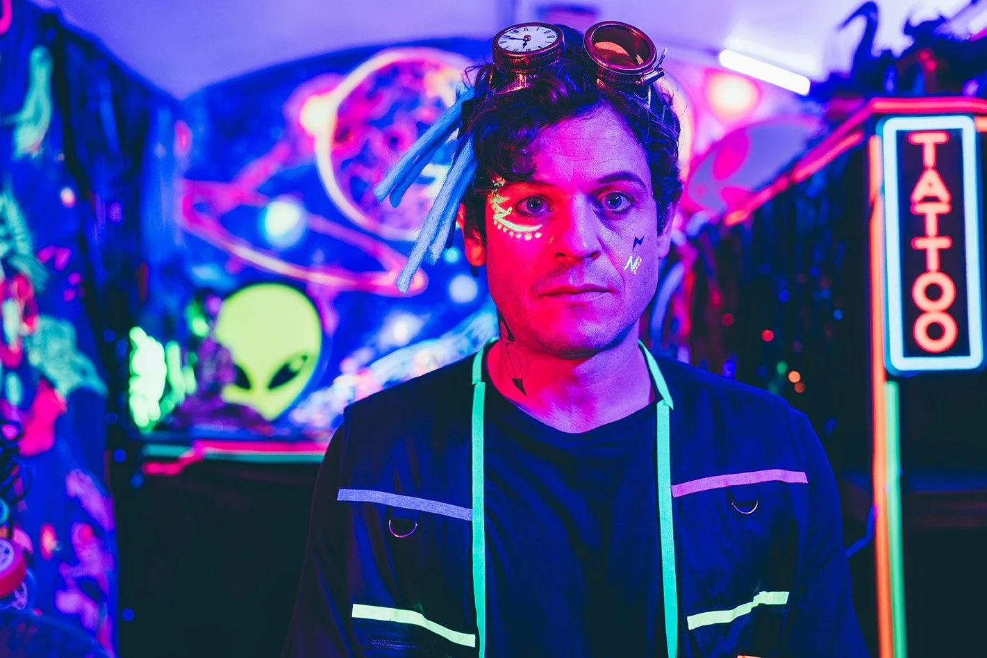A young man with curly hair wearing sunglasses on his head, face painted with neon designs, standing in front of a colorful neon-lit backdrop with an alien face and a tattoo shop sign.