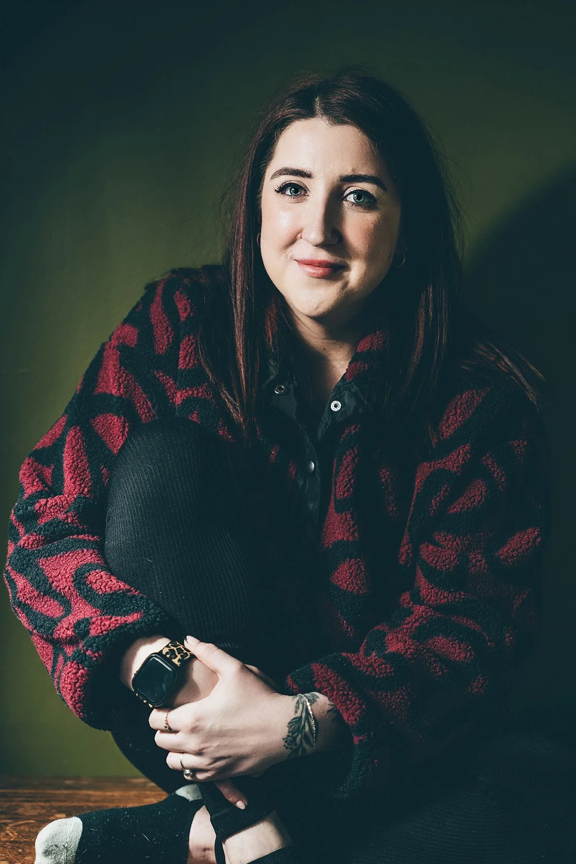 A woman with long dark hair and light skin is sitting on the floor against a green background, wearing a red and black patterned sweater, black pants, and a smartwatch. She is smiling slightly and has a tattoo on her wrist.