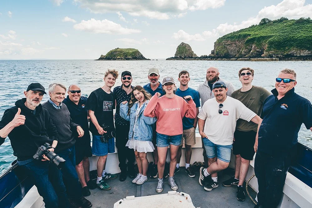 Group of 13 people posing on a boat with ocean and rocky islands in the background.