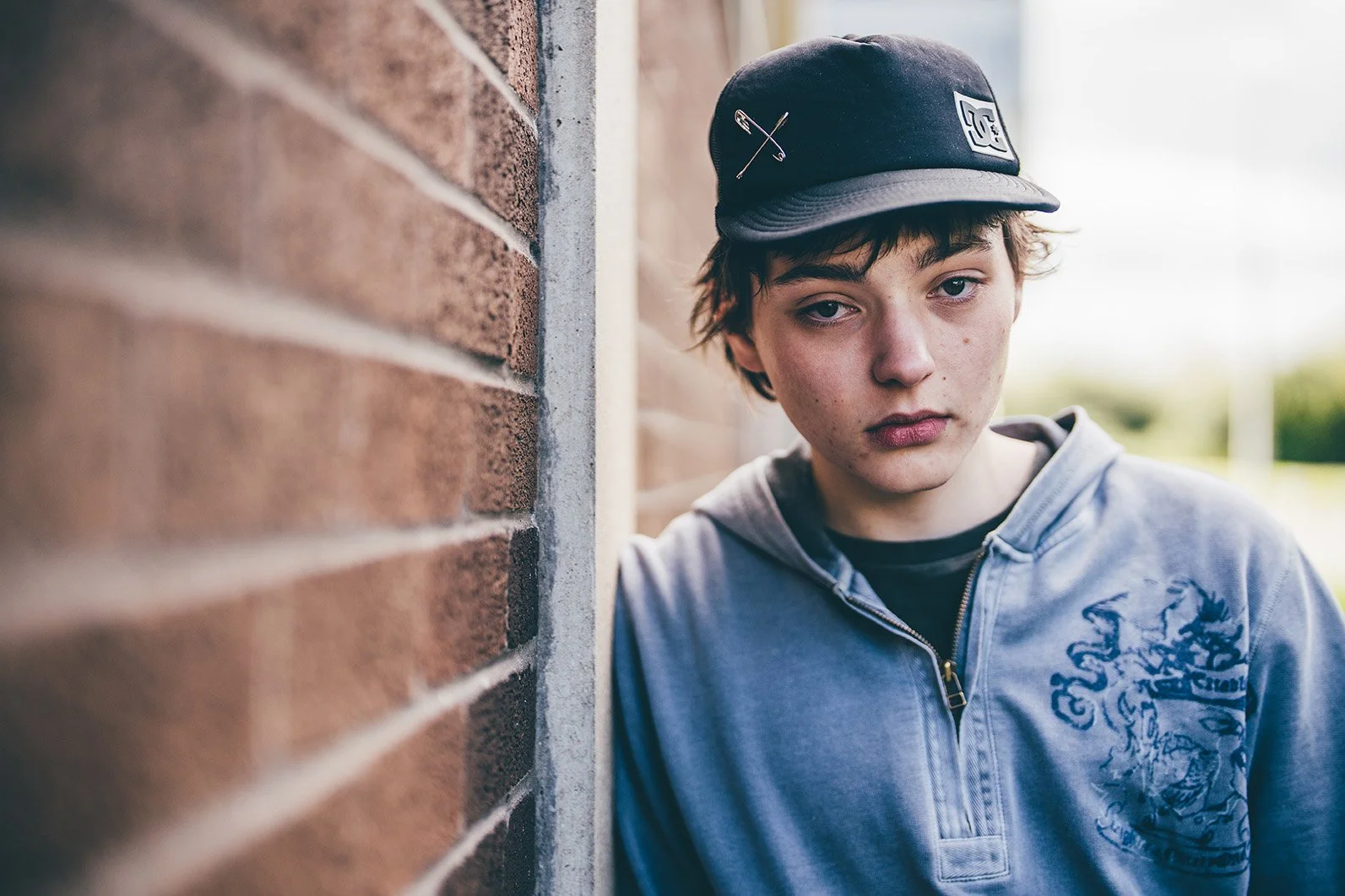 A teenage boy with light skin, messy brown hair, and a serious expression, standing next to a brick wall outdoors.
