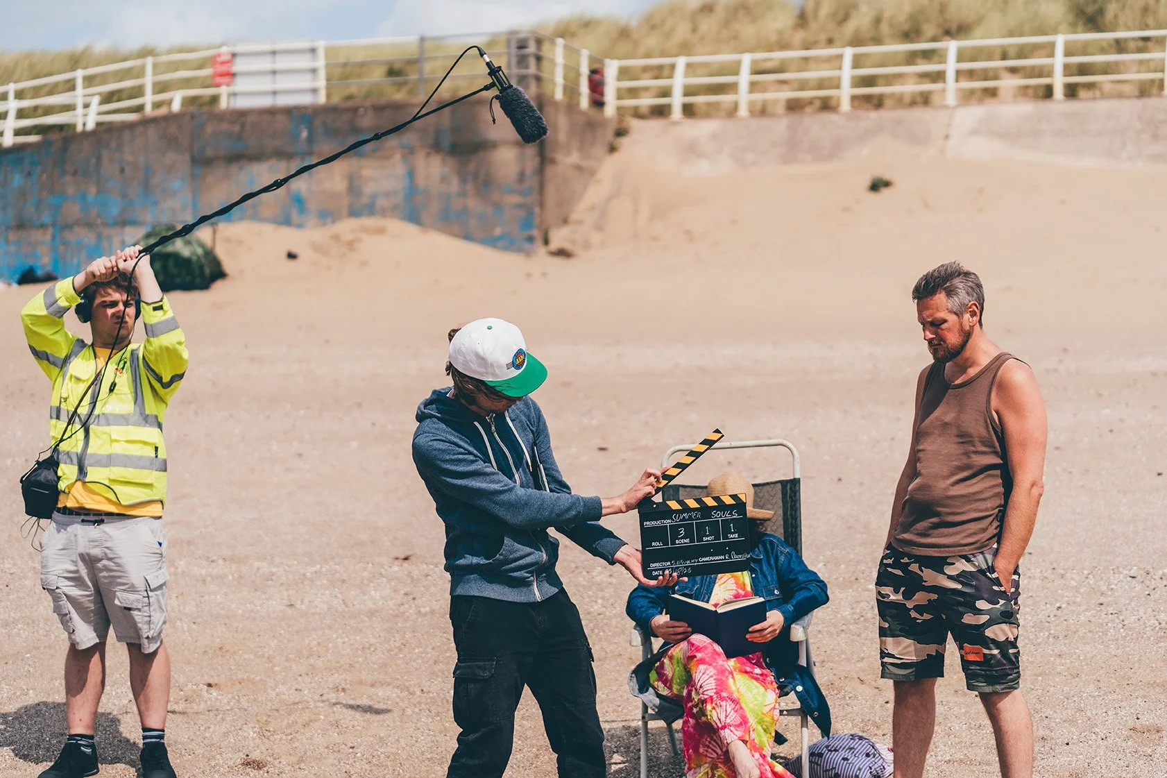 Behind the scenes of a film or video shoot on a sandy beach. There is a person holding a boom microphone, a crew member with a clapperboard, a man in a tank top and camo shorts, and a woman sitting in a beach chair reading a book.