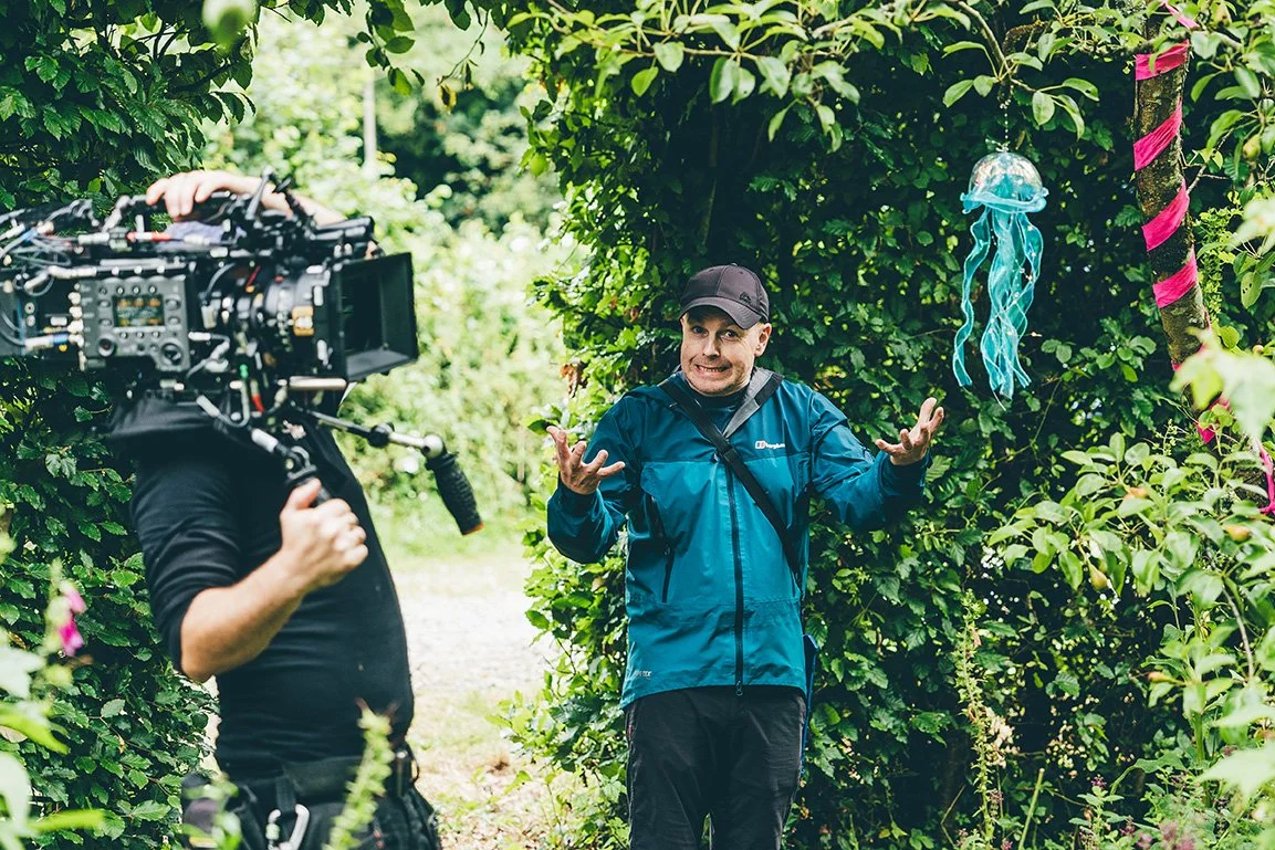 A man in outdoor clothing is talking to a camera crew in a lush garden with greenery and a jellyfish decoration hanging from a tree.