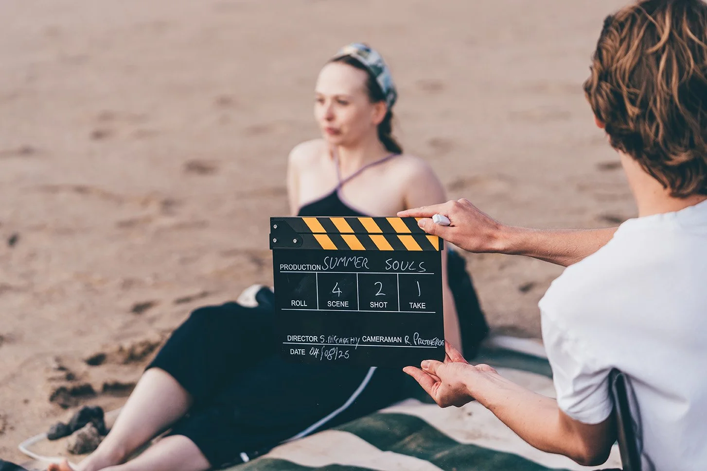 A film crew on a beach shooting a scene with a woman in a black dress, holding a clapperboard with the title 'Summer Souls'.