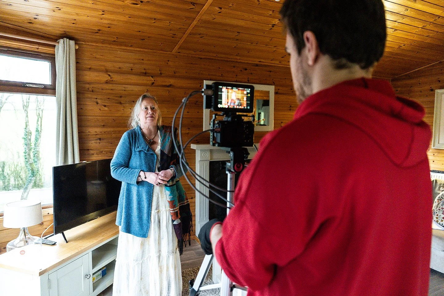 A woman standing in a room with wooden walls, being filmed by a man using a camera mounted on a stabilizer. The room has a large window with curtains, a TV on a white cabinet, and a mirror on the wall.