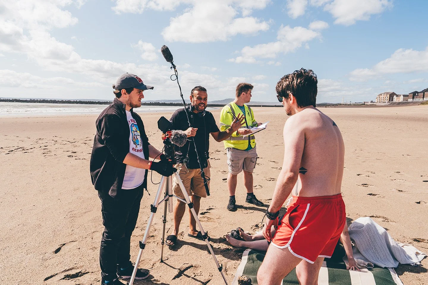 Filming crew interviewing a man in red shorts on a beach with a woman on the ground.