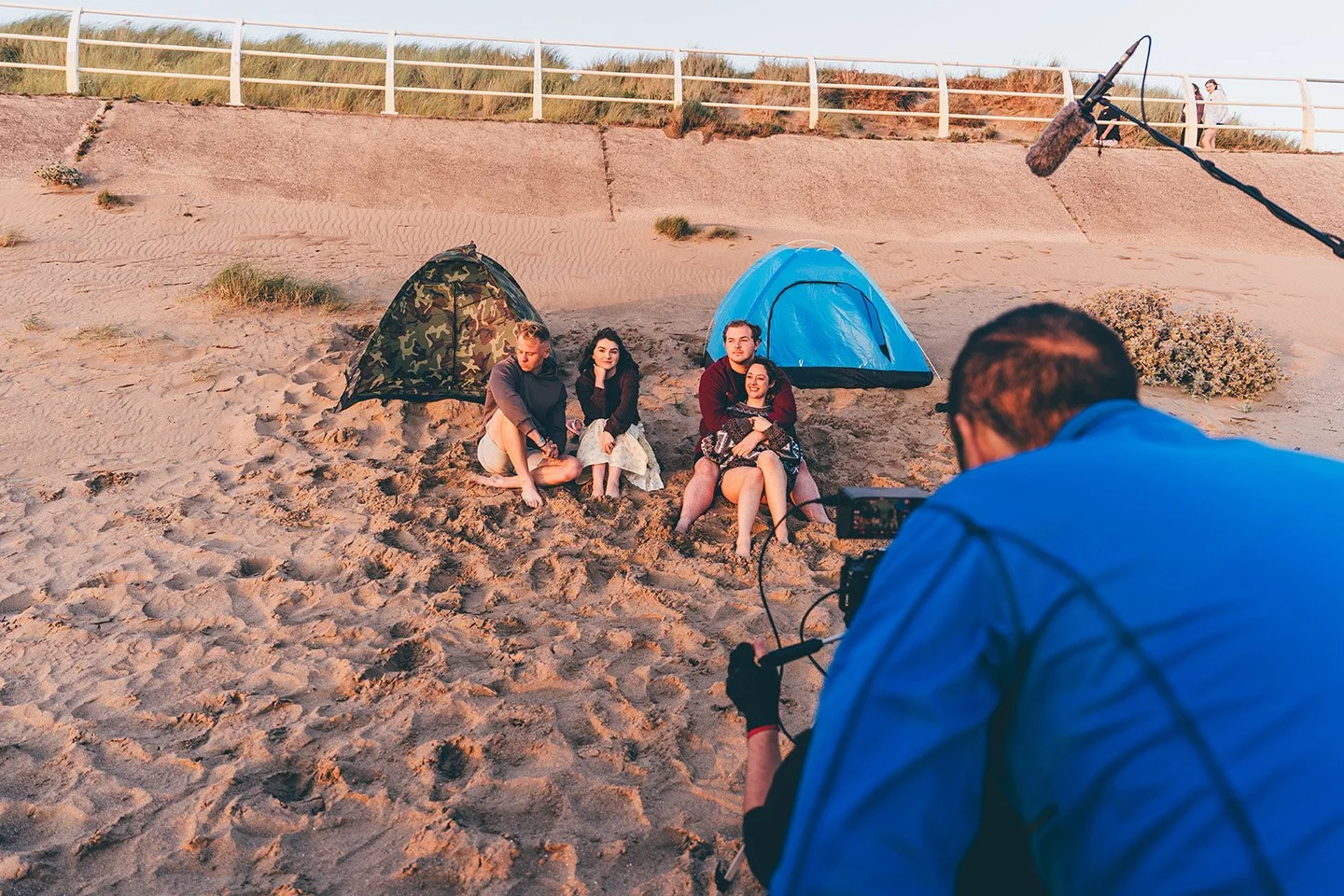 Film crew filming a scene with four actors sitting on the sand near tents on a beach.