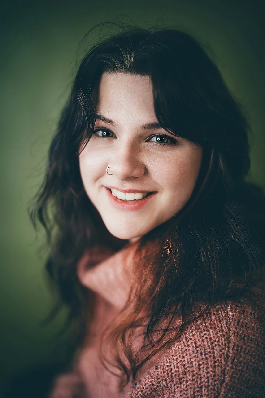 A smiling woman with dark wavy hair, blue eyes, and a nose piercing, wearing a pink knitted sweater.
