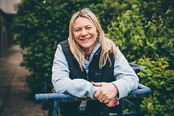 A woman with blonde hair smiling, leaning on a blue railing outdoors with greenery in the background.