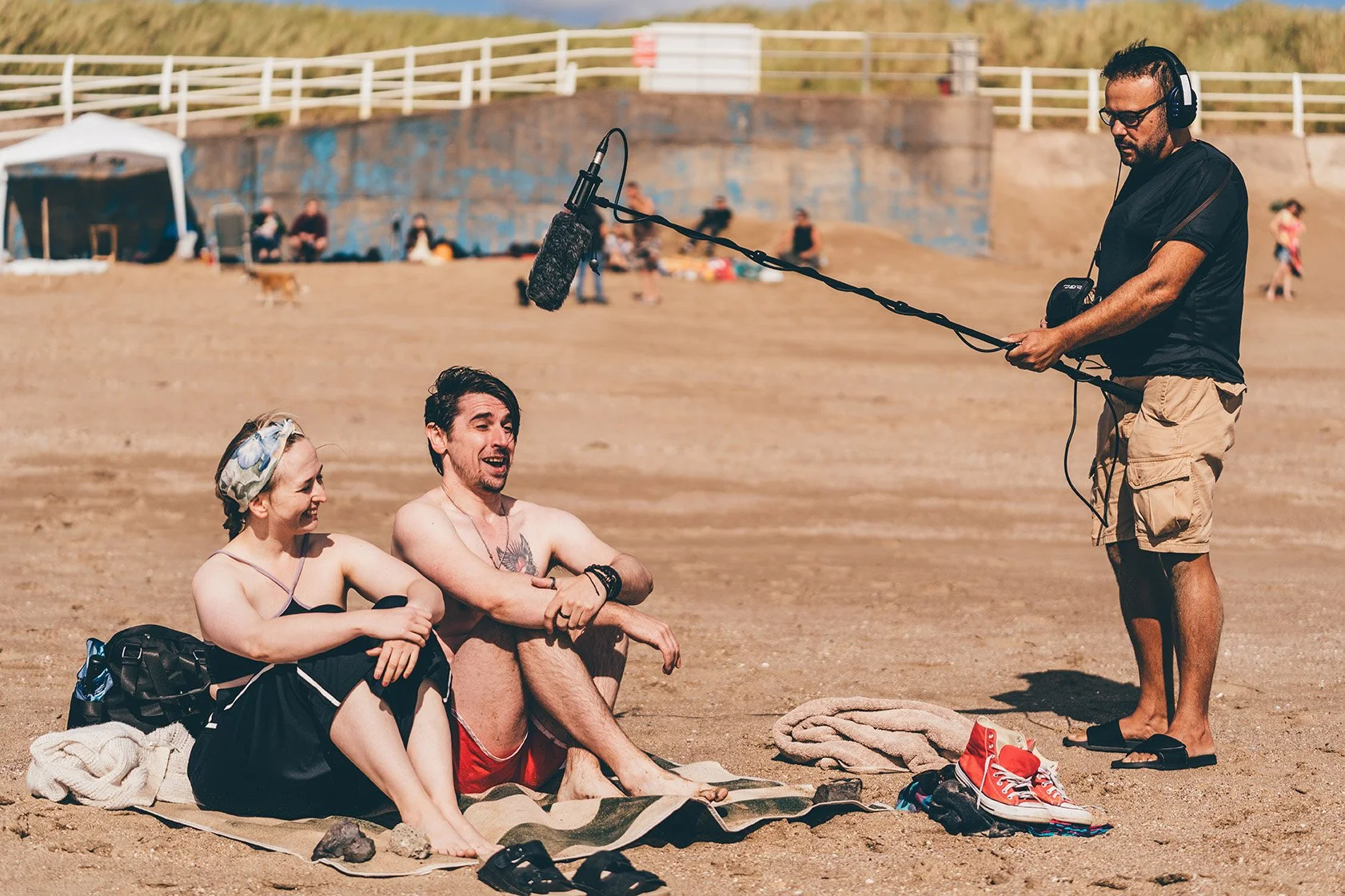 Actors on a beach set filming a scene with a sound technician recording sound.