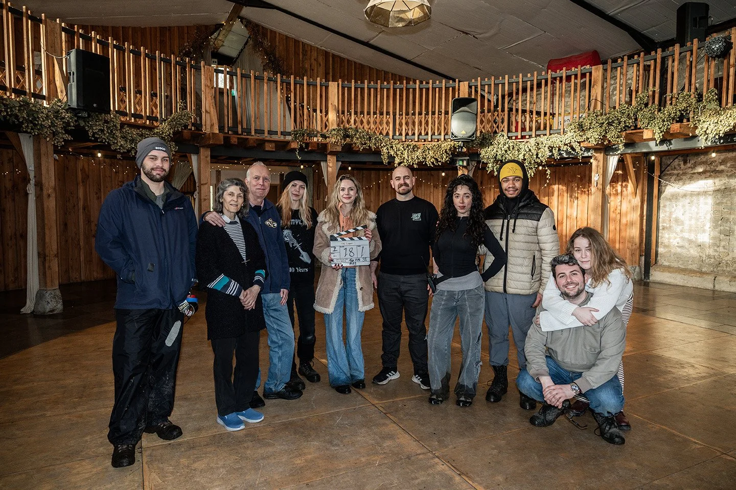 Group of ten people standing in a wooden indoor venue, smiling for a photo, with some holding a film clapperboard, suggesting a movie shoot or celebration event.