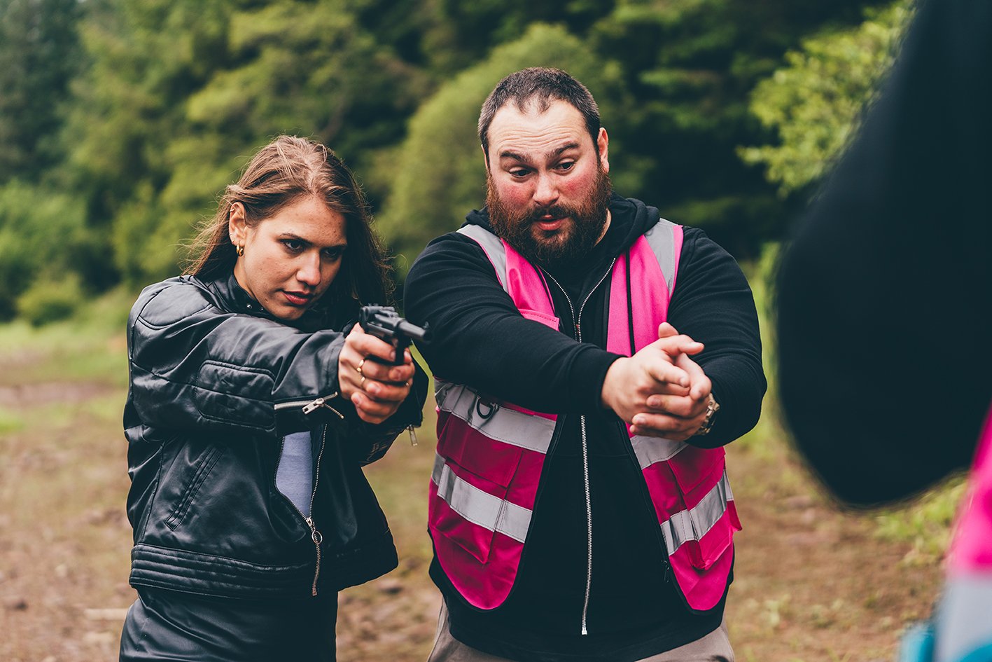 A woman with long brown hair aiming a handgun at a target, accompanied by a man with a beard and short hair observing, outdoors in a green, wooded area.