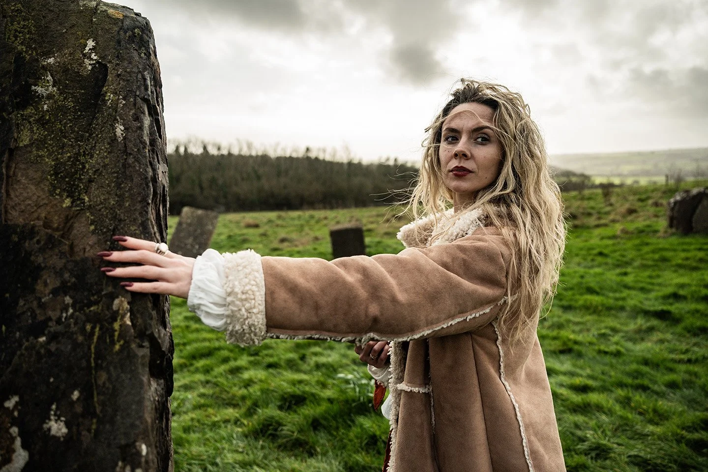 A woman with long curly blonde hair and wearing a tan coat with shearling lining stands outdoors in a grassy field, touching a moss-covered stone or tree trunk, with a cloudy sky in the background.