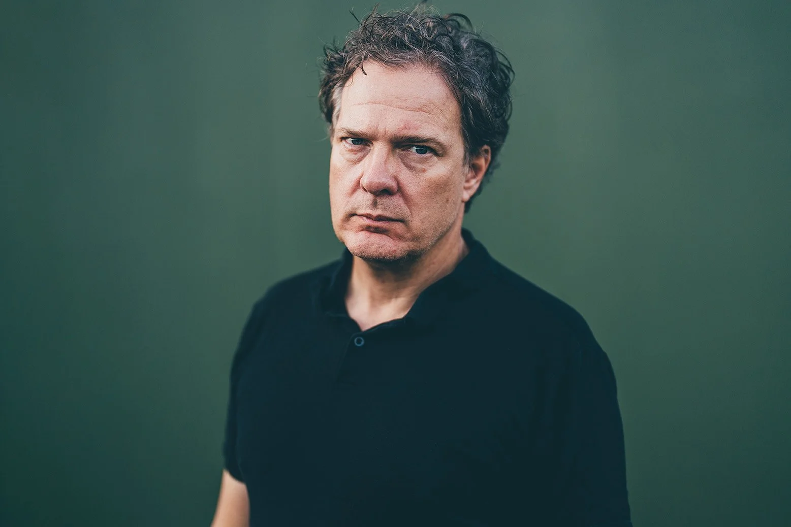 A middle-aged man with disheveled dark hair and a serious expression looking directly at the camera. He is wearing a black shirt and is standing against a plain, muted green background.