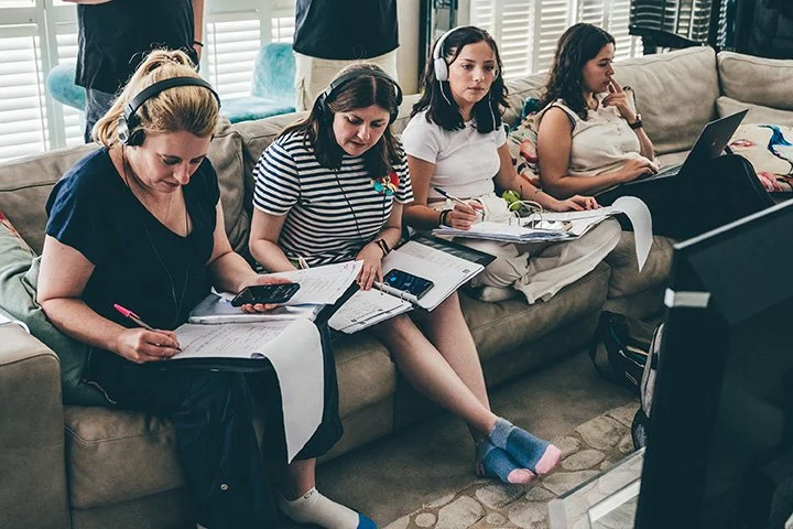 Four women sitting on a couch, wearing headphones, working with notebooks, papers, and electronic devices during a recording or broadcasting session.