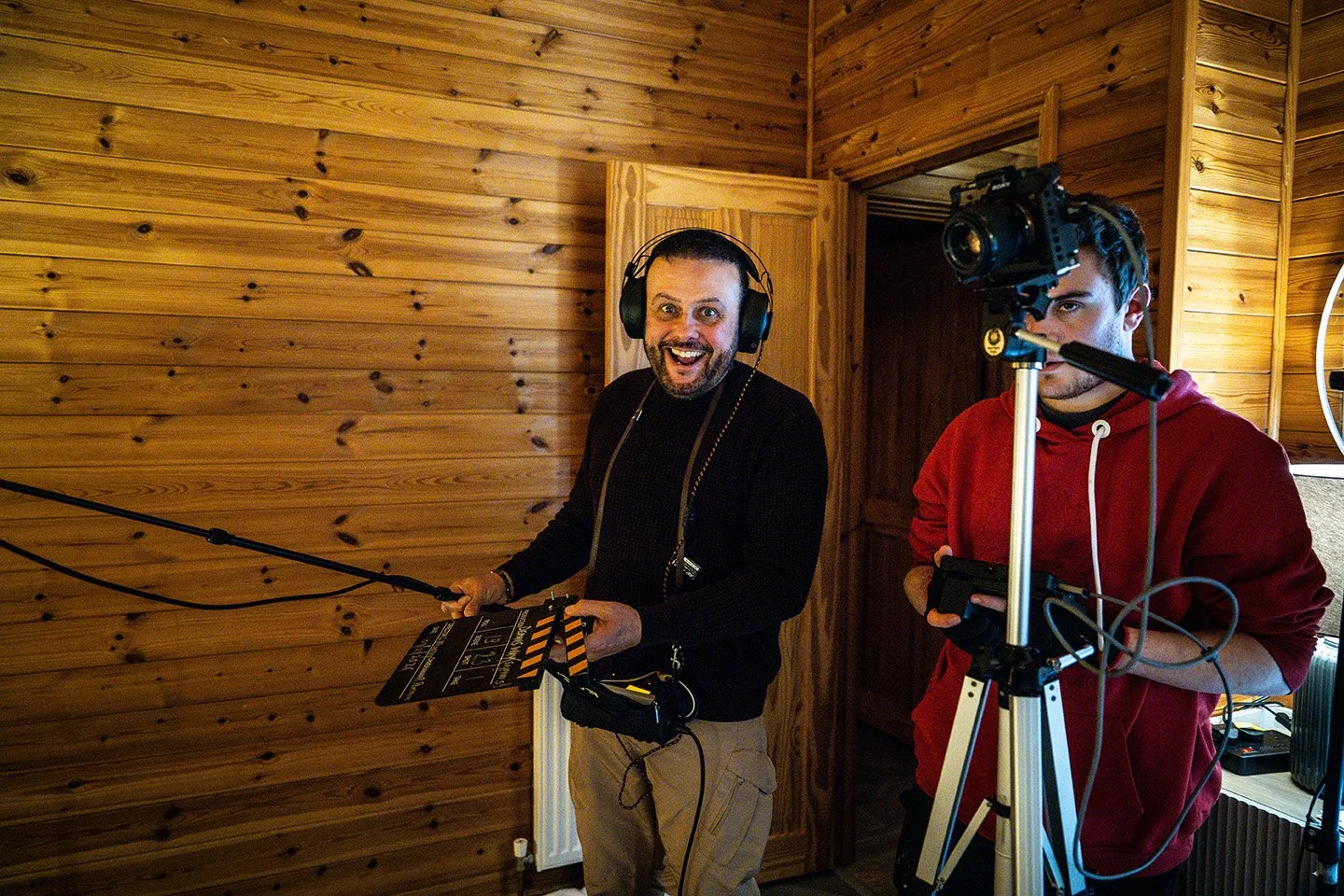 Two men working on a film set inside a wood-paneled room, one with a clapboard and headphones, the other operating a camera on a tripod.