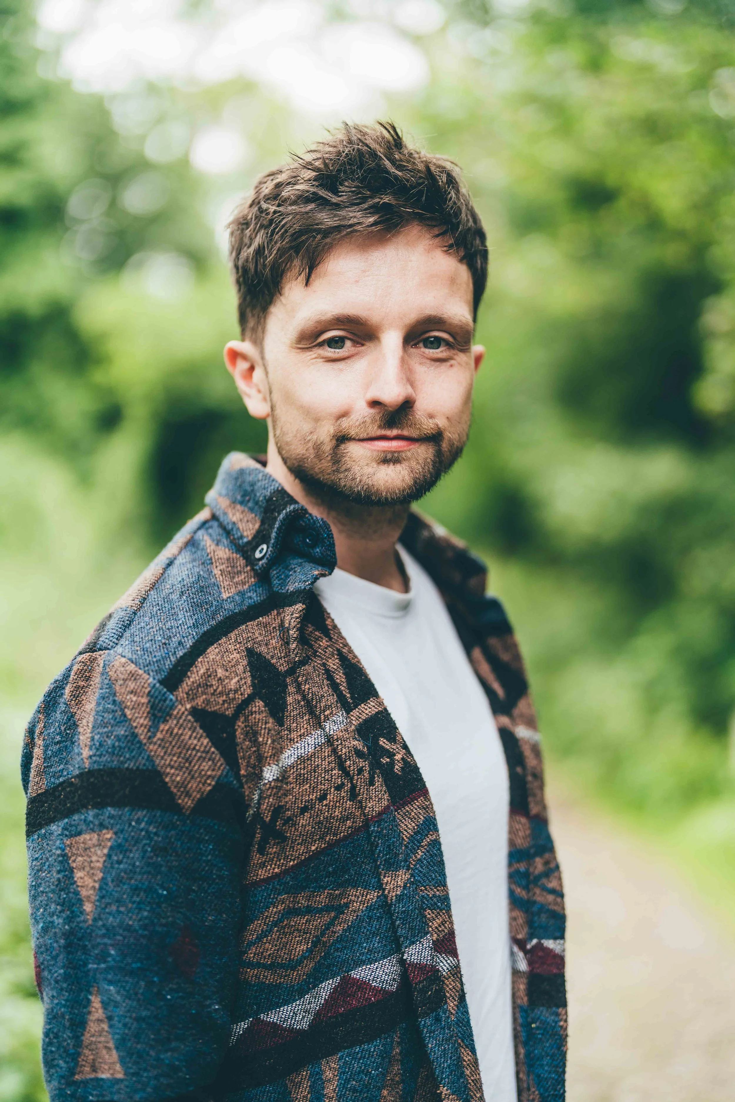 A man with brown hair and a beard, wearing a colorful patterned jacket over a white T-shirt, standing outdoors with blurred greenery in the background.