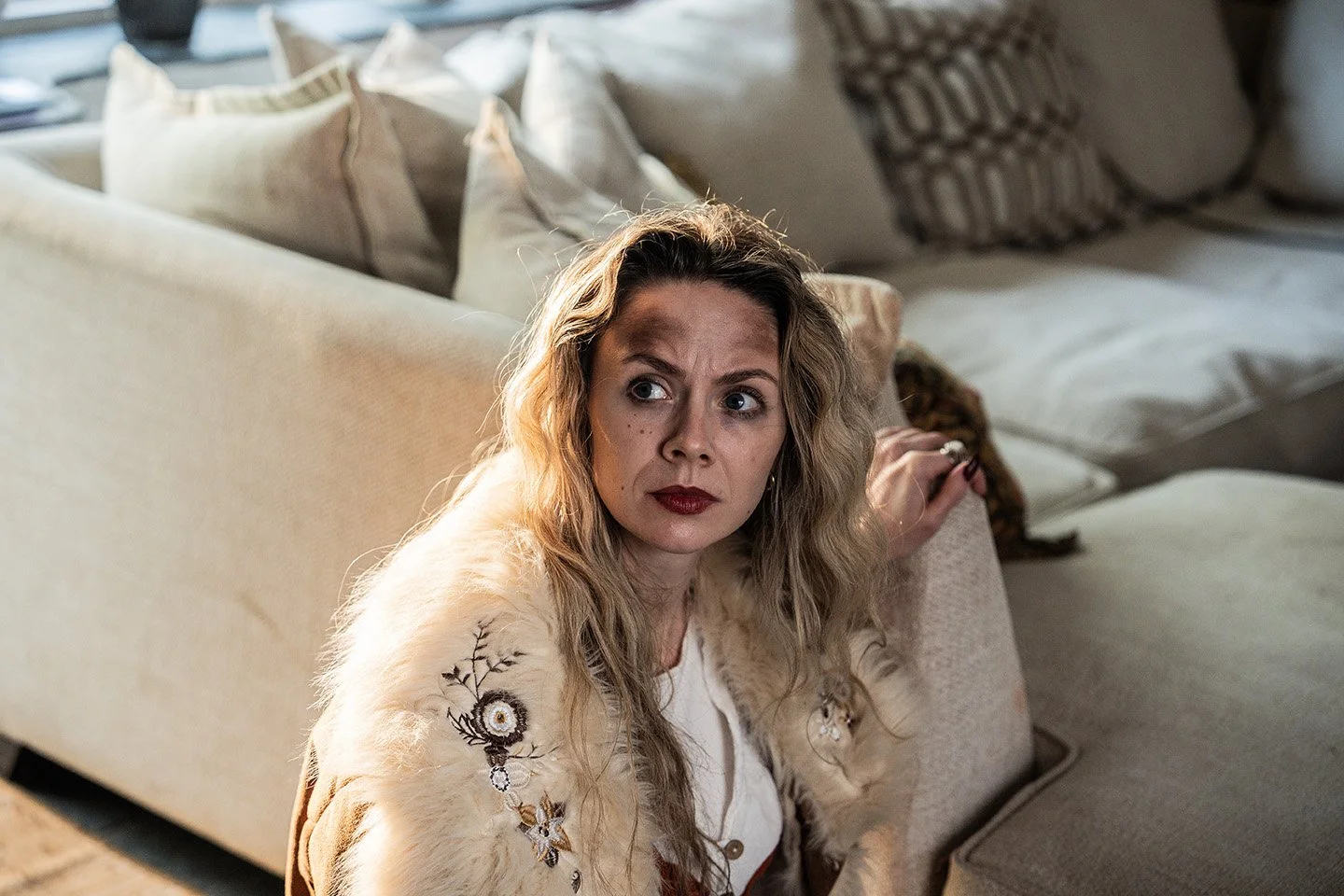 A woman with wavy blonde hair sitting on a couch, looking to her right with a serious expression. She is wearing a beige, fur-lined jacket with embroidered floral designs on the sleeve. Behind her are multiple pillows on the couch.